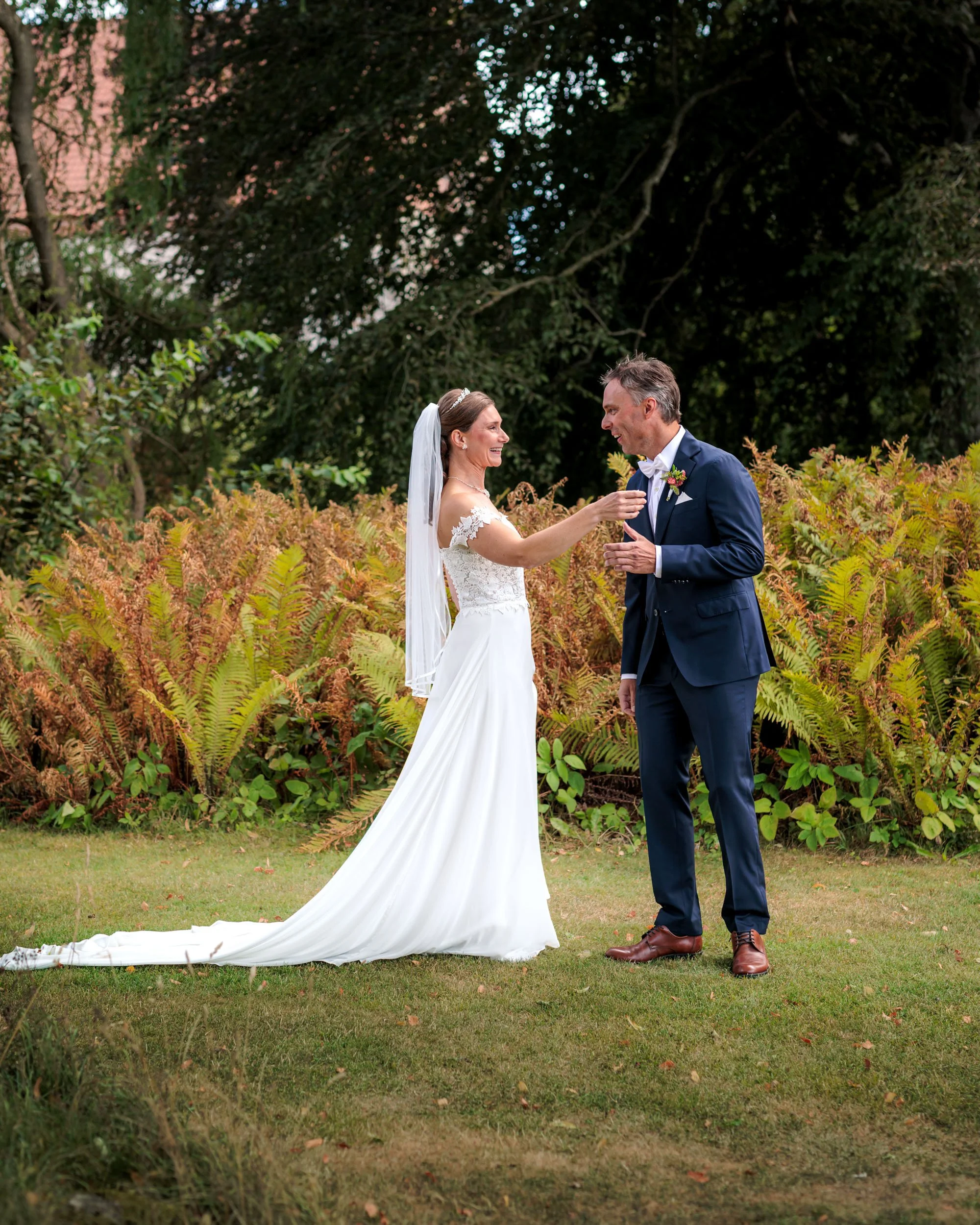 En brud och en brudgum står utomhus på en gräsmatta, omgivna av buskar och träd i bakgrunden. De ler mot varandra, och bruden räcker fram en hand mot brudgummen, som är klädd i en mörkblå kostym. Det är en lycklig och romantisk stund, troligen under 