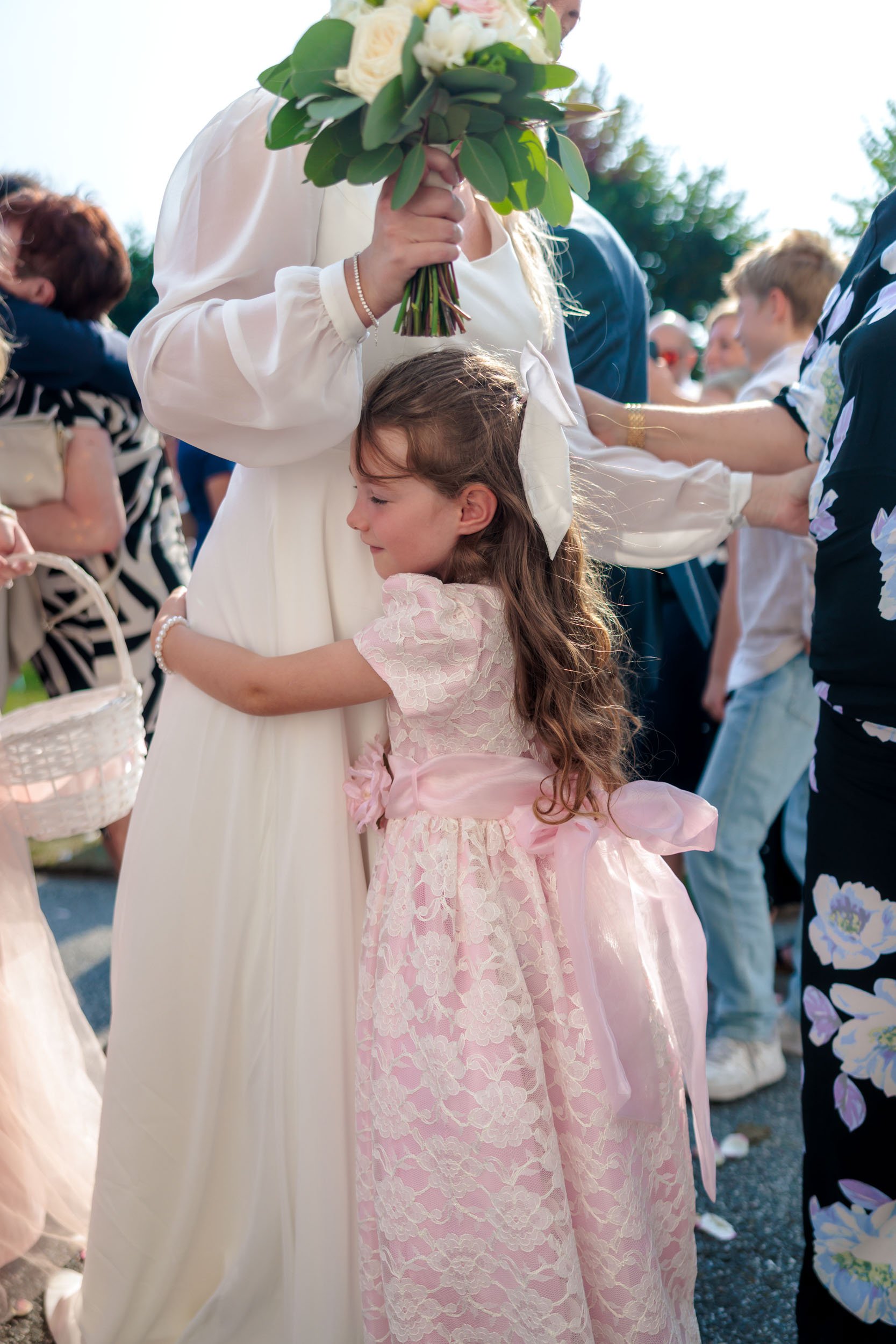 En ung flicka i rosa klänning omfamnar en kvinna i vit klänning på ett utomhusbröllop.
