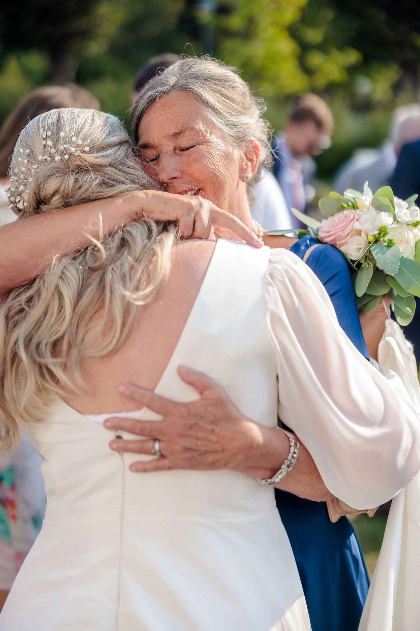 Två kvinnor omfamnar varandra i en varm omfamning, en av dem håller en bukett blommor, och de verkar vara glada och känslosamma. Bakgrunden visar andra personer och gröna träd.