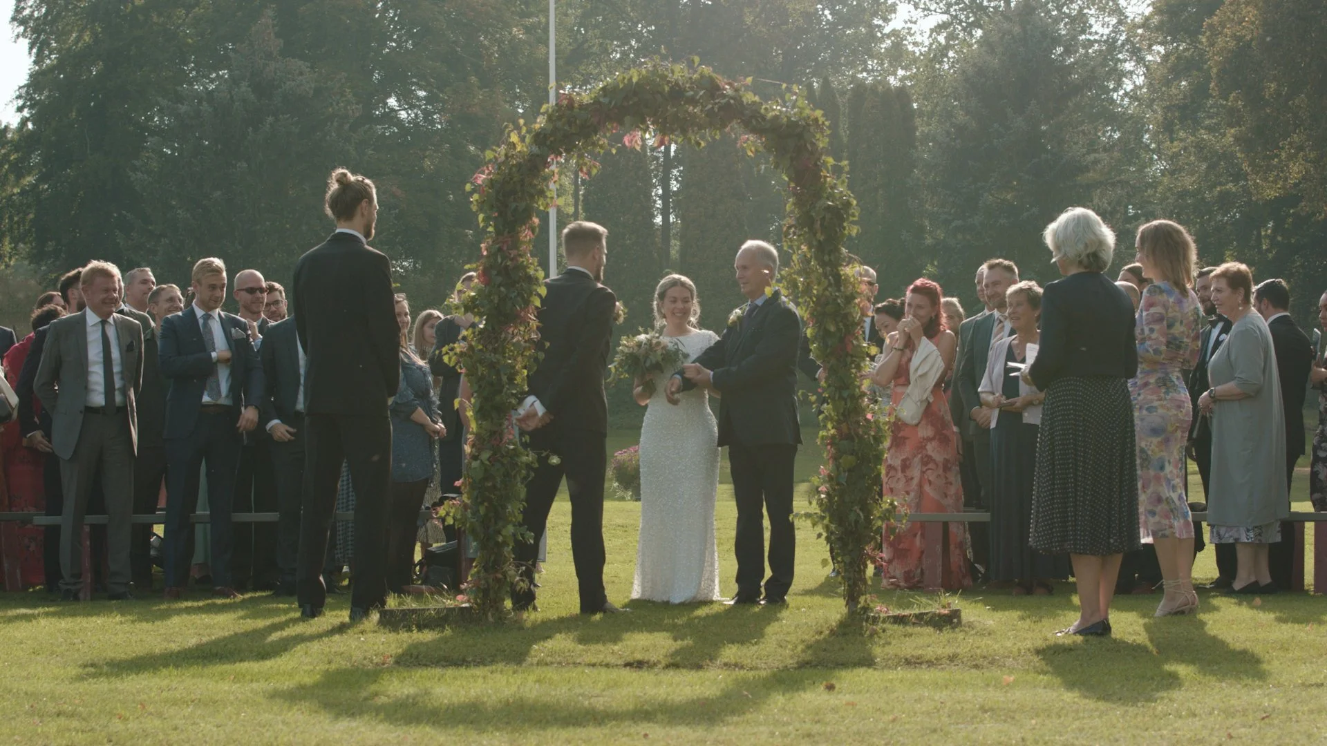 Bröllopspar under utomhusvigsel med blomsterbåge och gäster – vigsel Sverige