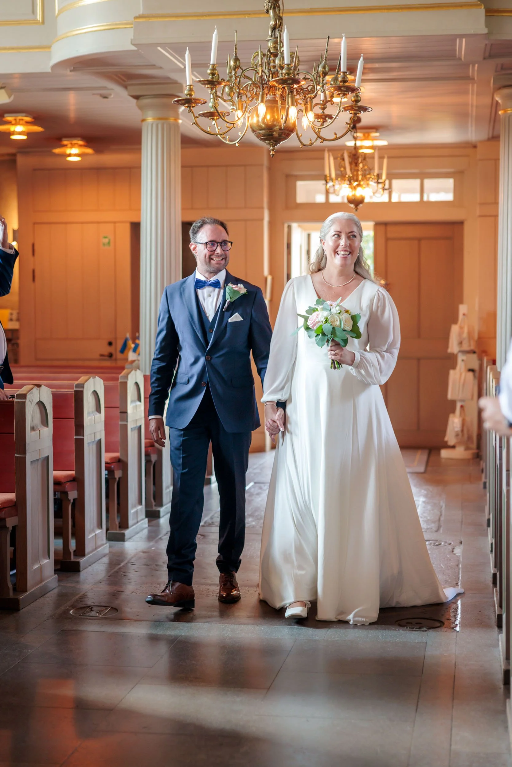 En nygift kvinna i vit brudklänning bär en bukett medan hon går med en man i mörk kostym, i en festlig lokal med kristallkronor.