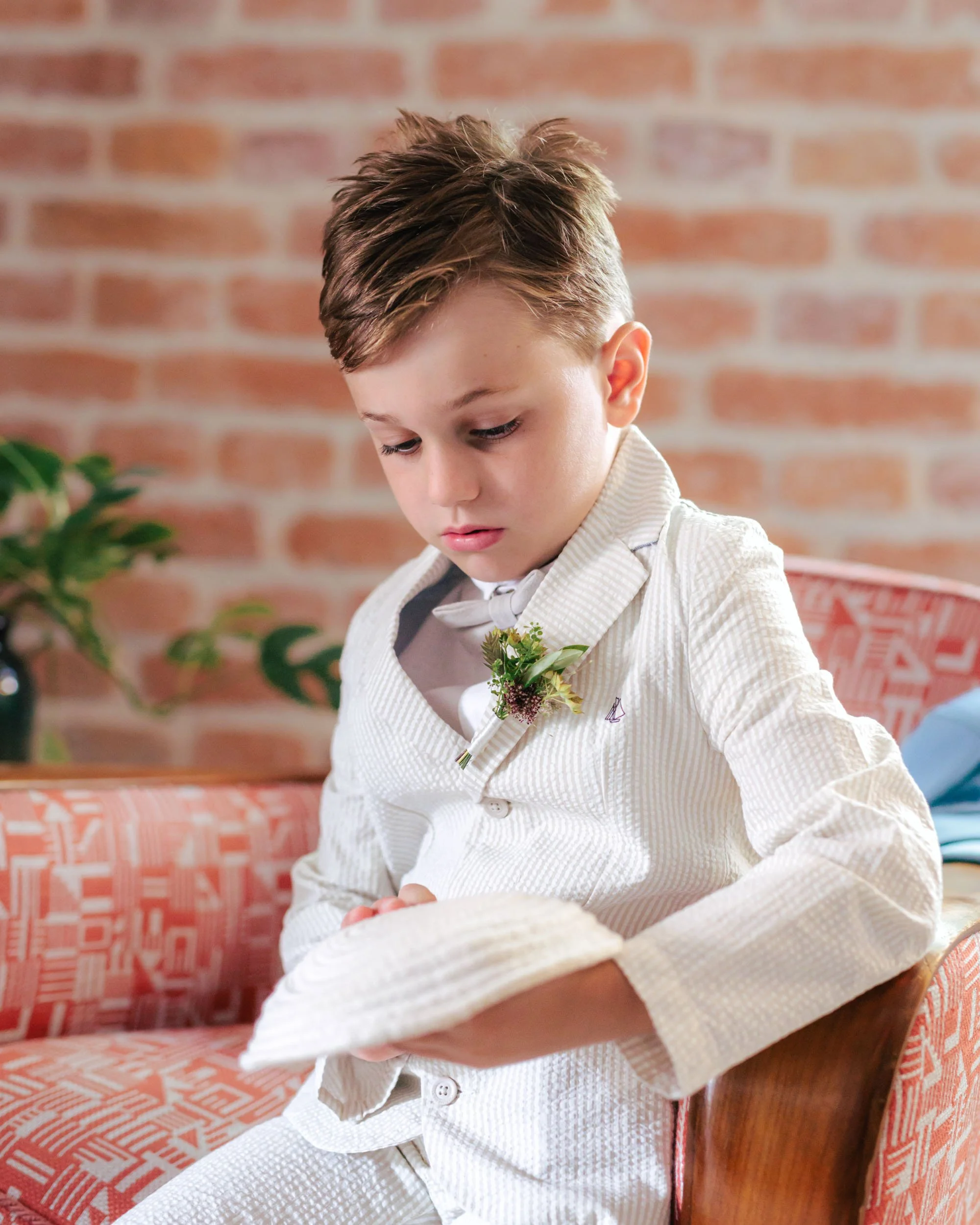 En pojke i vit bröstjacka med blomsterklädd knapphål sitter på en rödmönstrad soffa, tittar ner.