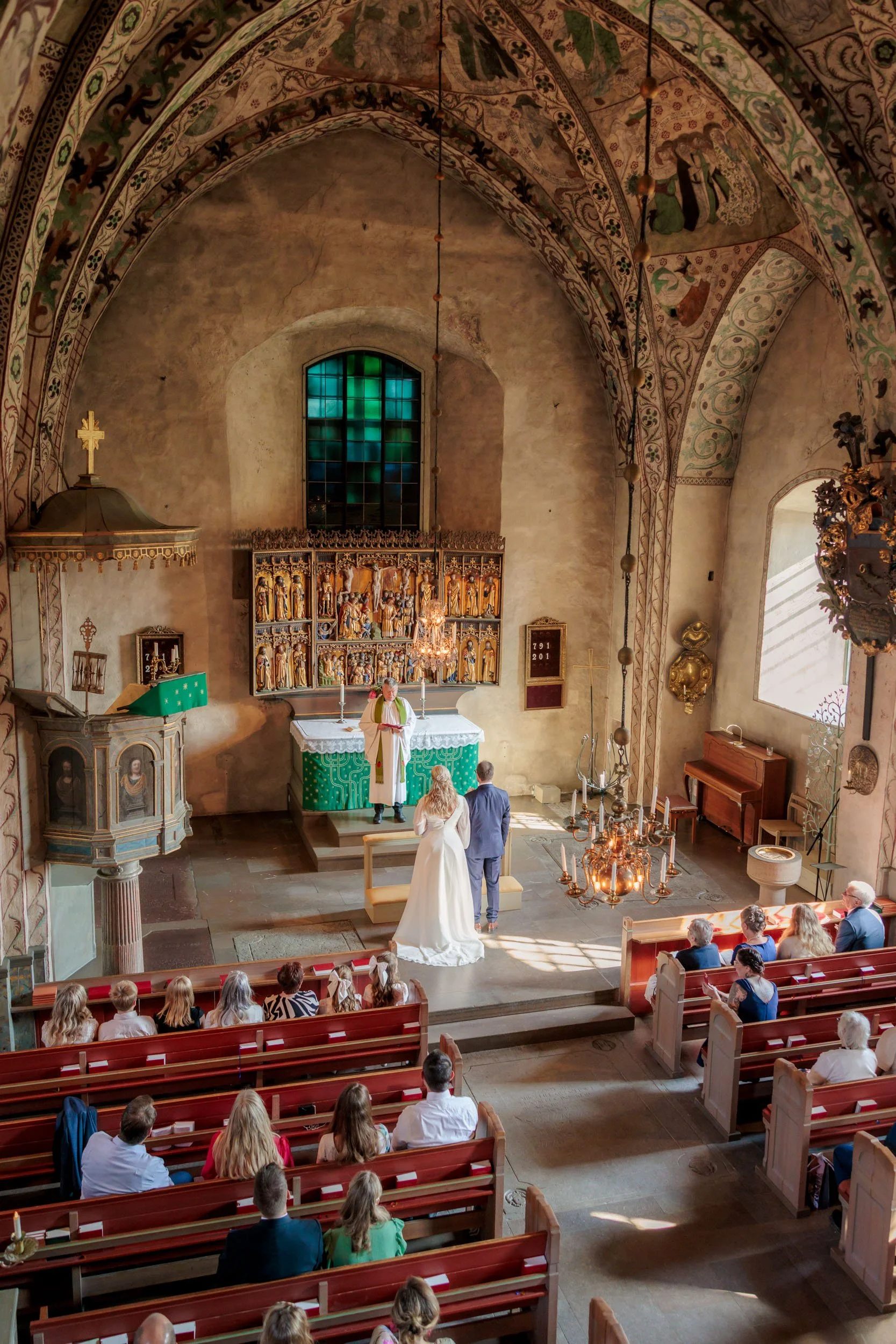 En brud och en man står framför en präst i en kyrka under ett bröllop. Klockor och gudstjänstföremål finns i kyrkan, och gäster sitter i bänkar.
