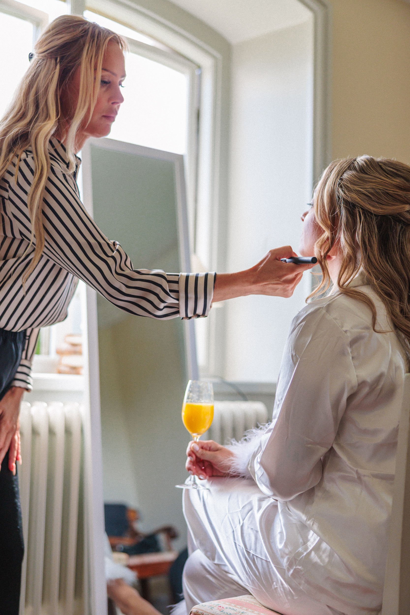 En kvinna i svart och vit randig blus sminkar en annan kvinna med ljus hår, som sitter i en vit klänning och håller ett glas apelsinjuice.
