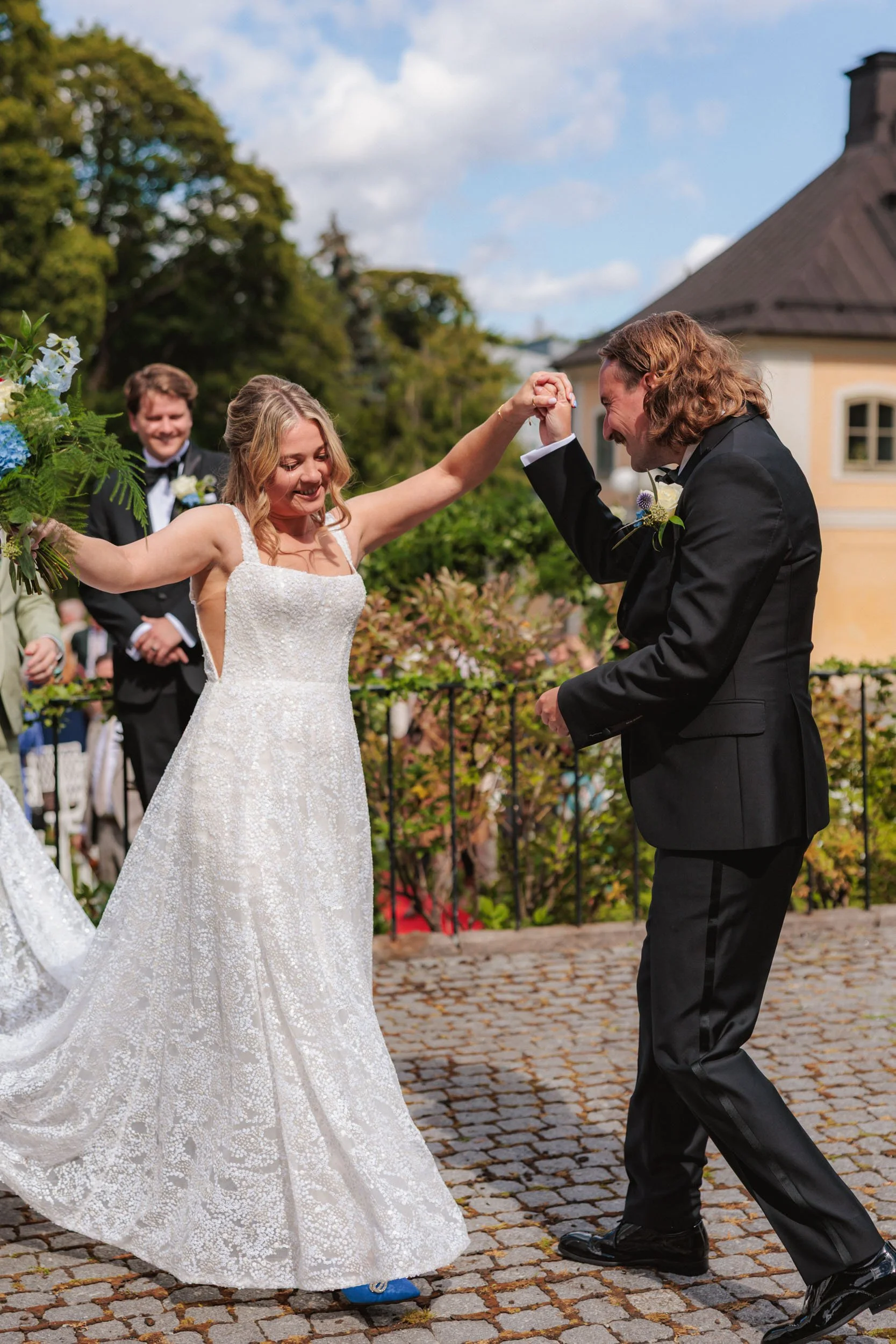 En brud och en brudgum dansar ute på en stenlagd plats under ett bröllop, med ett leende på läpparna. Bruden bär en vit, spetsklädd brudklänning och brudgummen en svart kostym.