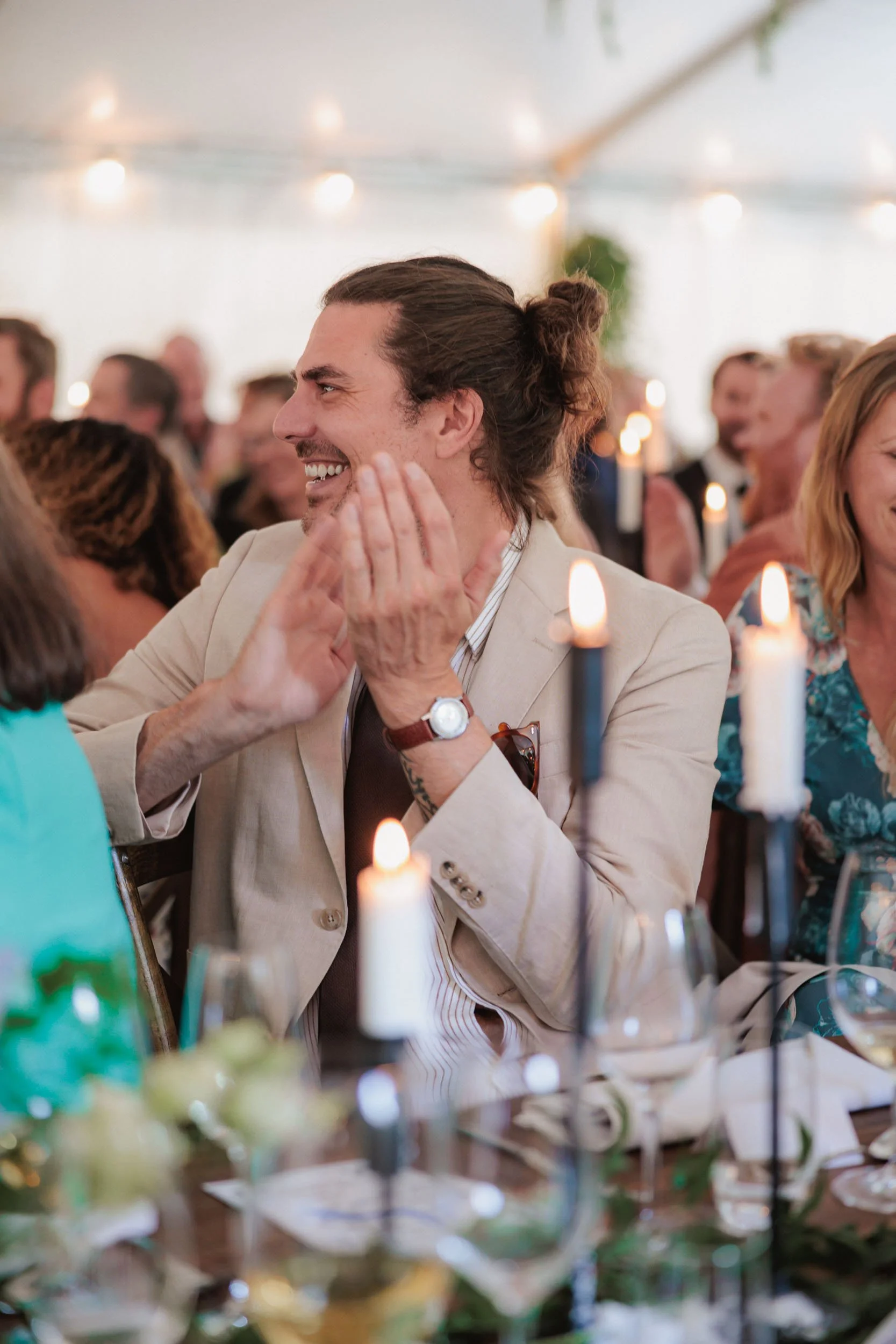 En man med långt hår i en knut, klädd i beige kavaj och vit skjorta, skrattar och talar med någon vid ett bord på ett festligt evenemang, omgiven av middagsgäster och ljuslyktor.