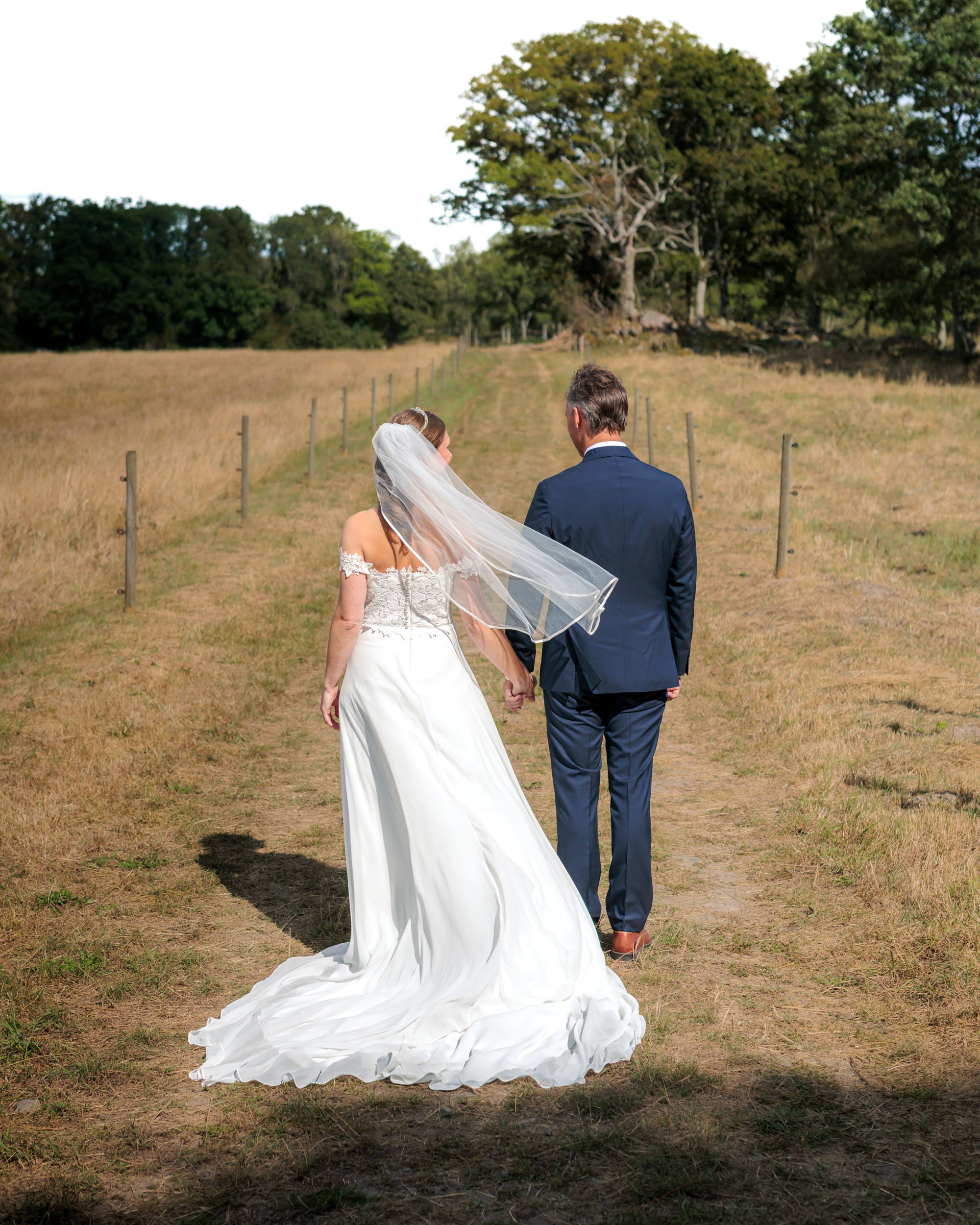 En brud och en bröllopsklänning håller hand och går på en bana i ett fält med träd i bakgrunden.