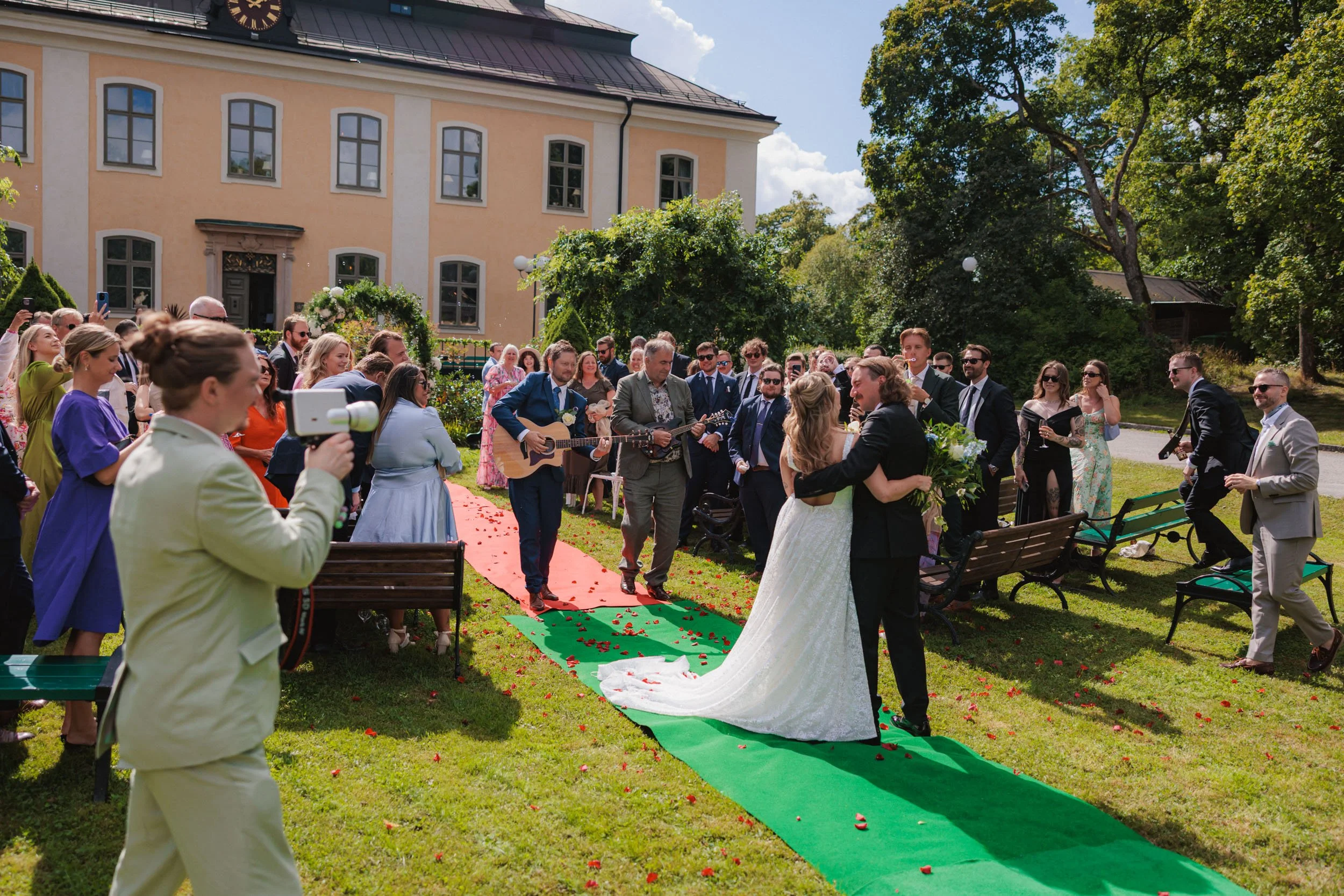 Bröllopsceremoni med nygifta par som kramas, omgivna av gäster utomhus på en solig dag.