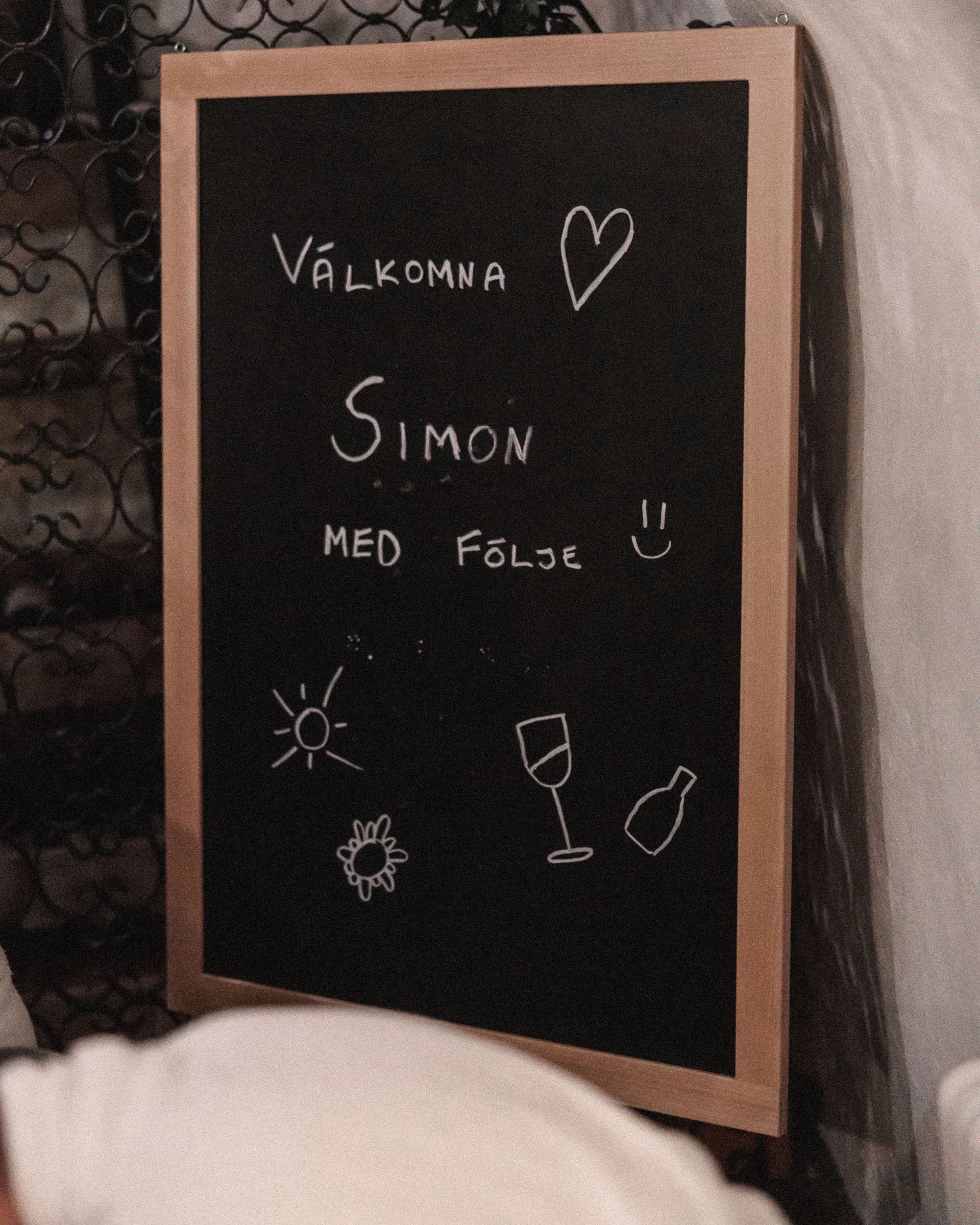 En svart griffeltavla med text på svenska och enkla teckningar av en sol, en blomma, ett vin- och en ölglas. Texten säger "Välkomna Simon med följje" med ett hjärta, ett leende, och glada ögon.
