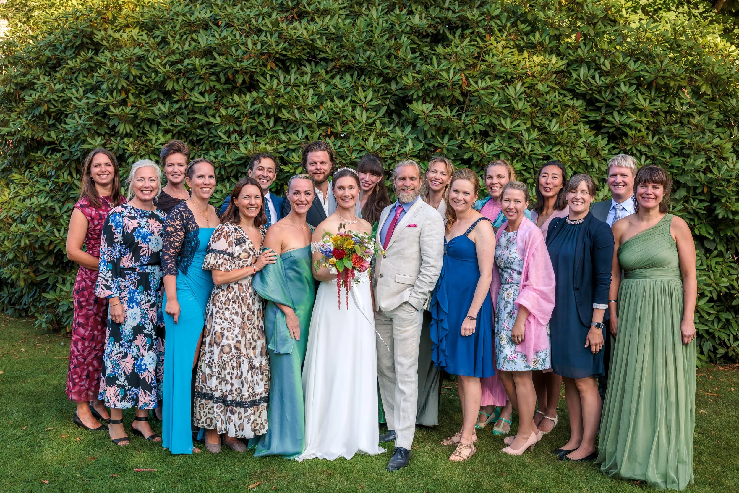 Grupp av människor på bröllop utomhus, alla ler, några håller i blommor, grön växtlighet i bakgrunden.