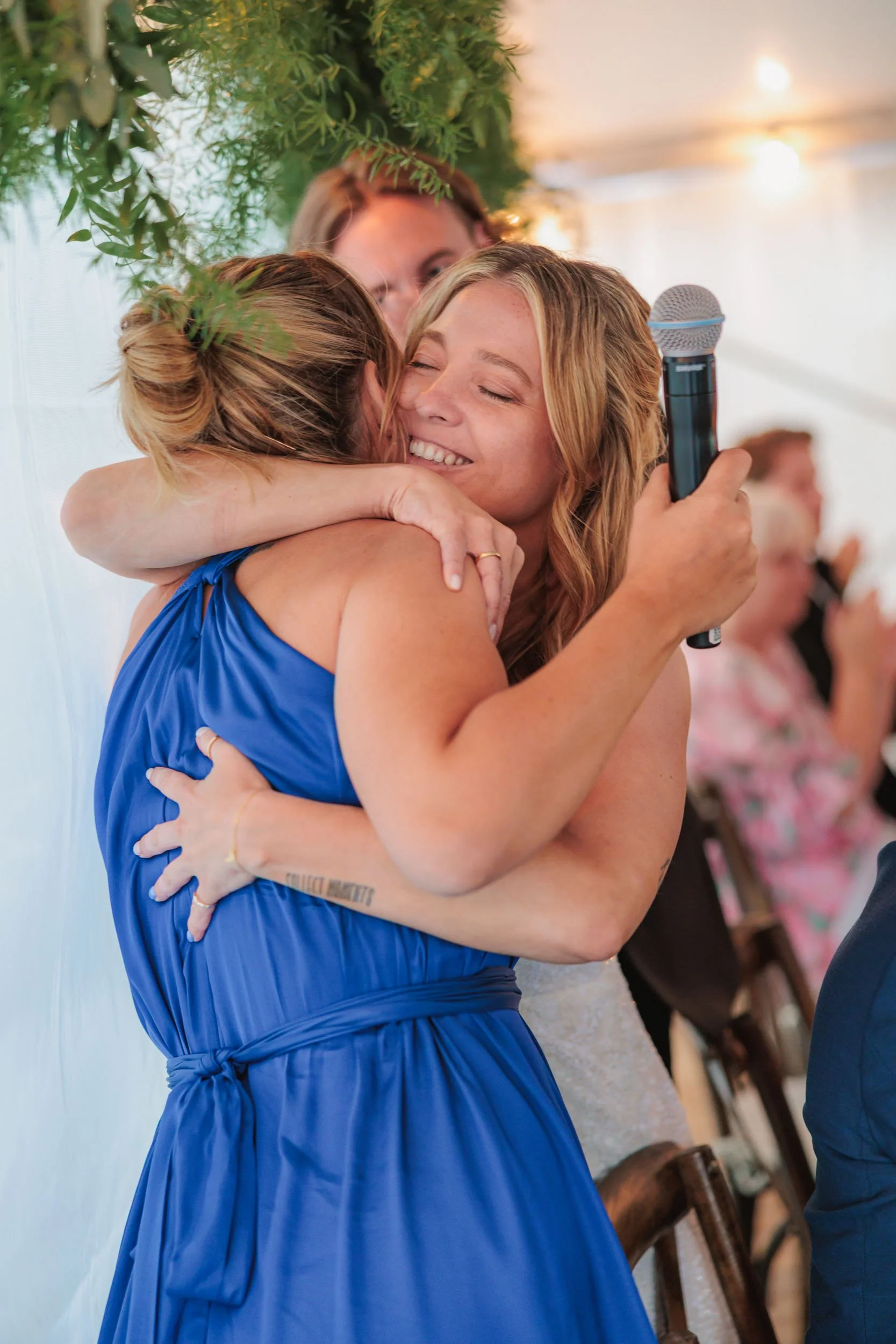 Två kvinnor omfamnar varandra på en fest, en av dem håller ett mikrofon och är glad.