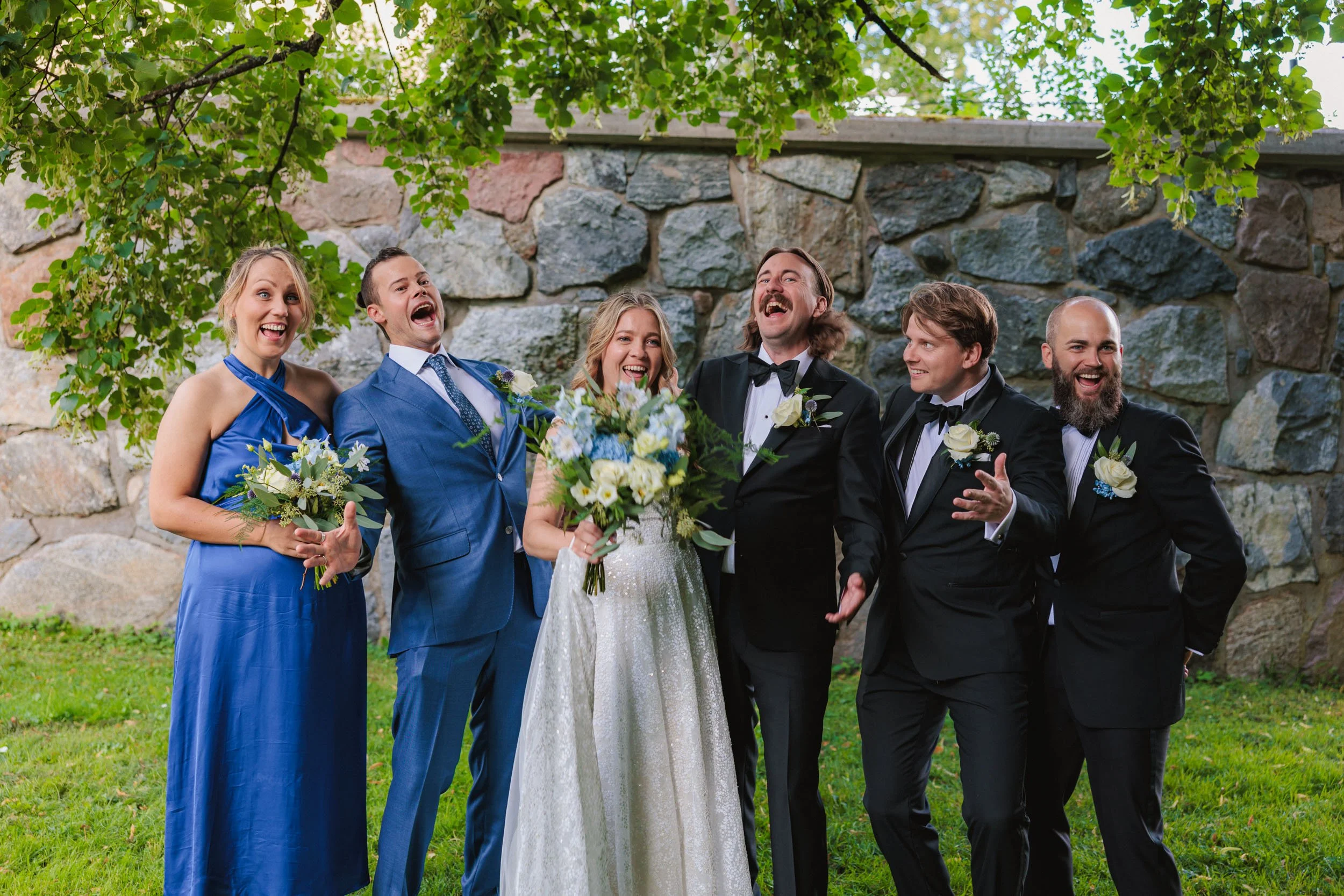 En grupp människor i formella kläder som skrattar och ler, troligen vid ett bröllop utomhus med en stenmur och grönt lövträd i bakgrunden.