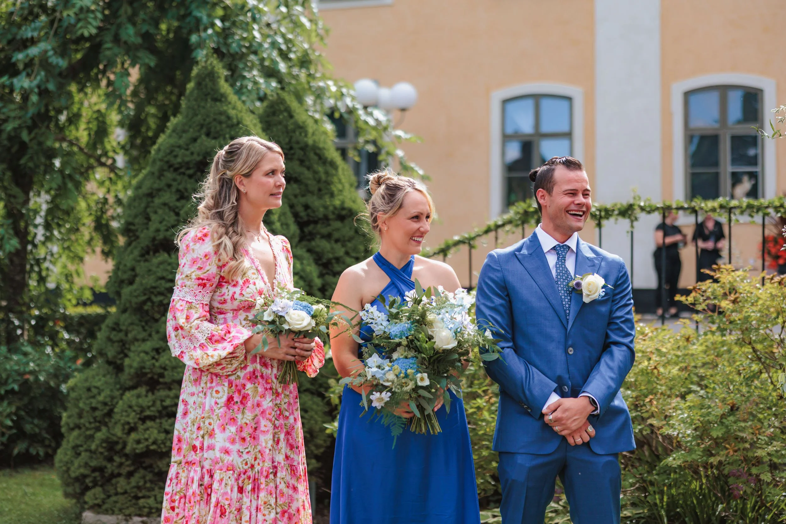 Tre personer står utomhus, två kvinnor i blå och rosa klänningar och en man i blå kostym, alla håller buketter, skrattar och deltar i ett bröllop eller ceremoni, med ett hus och ornamenterade trädgårdar i bakgrunden.