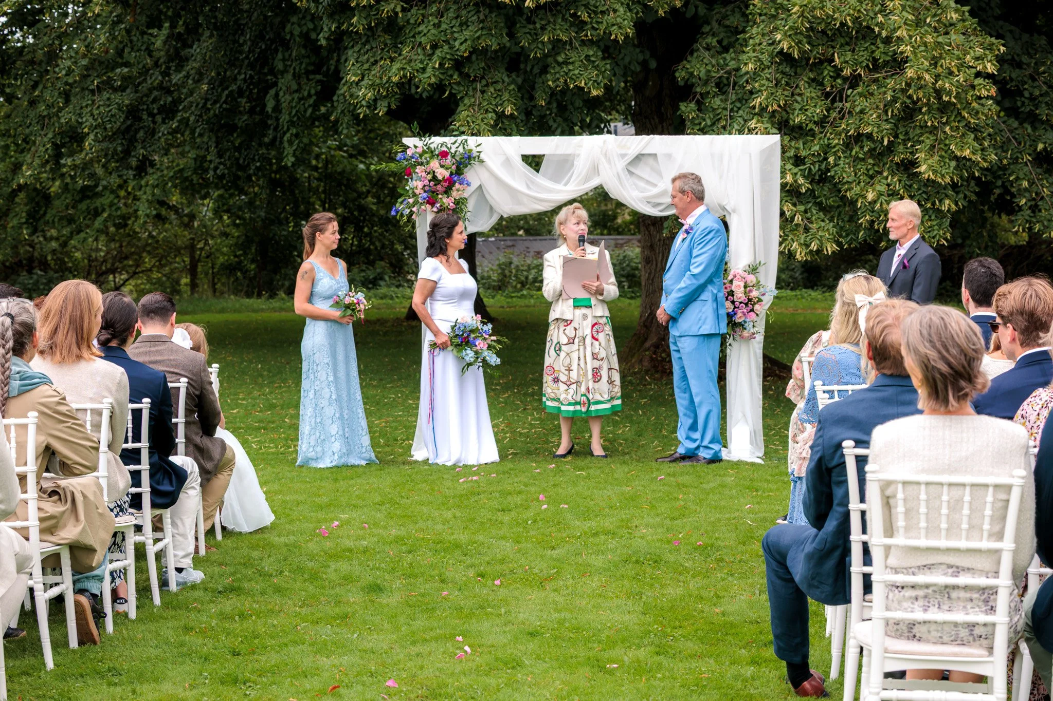Vigsel utomhus i grön trädgård med vit blomsterbåge och sittande gäster