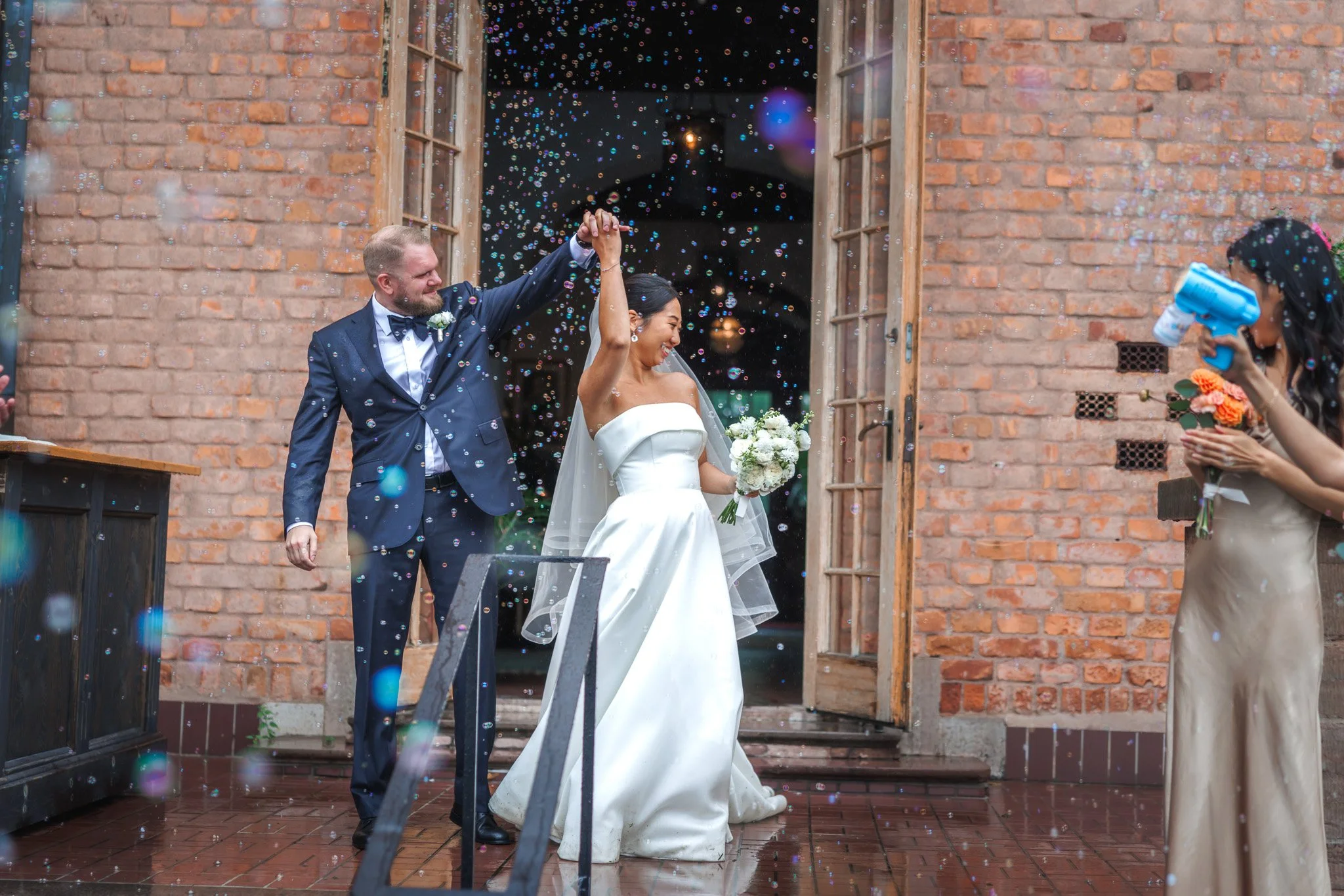 Brudpar dansar ut ur tegelbyggnad omgivet av såpbubblor i regn med vit bukett och smoking