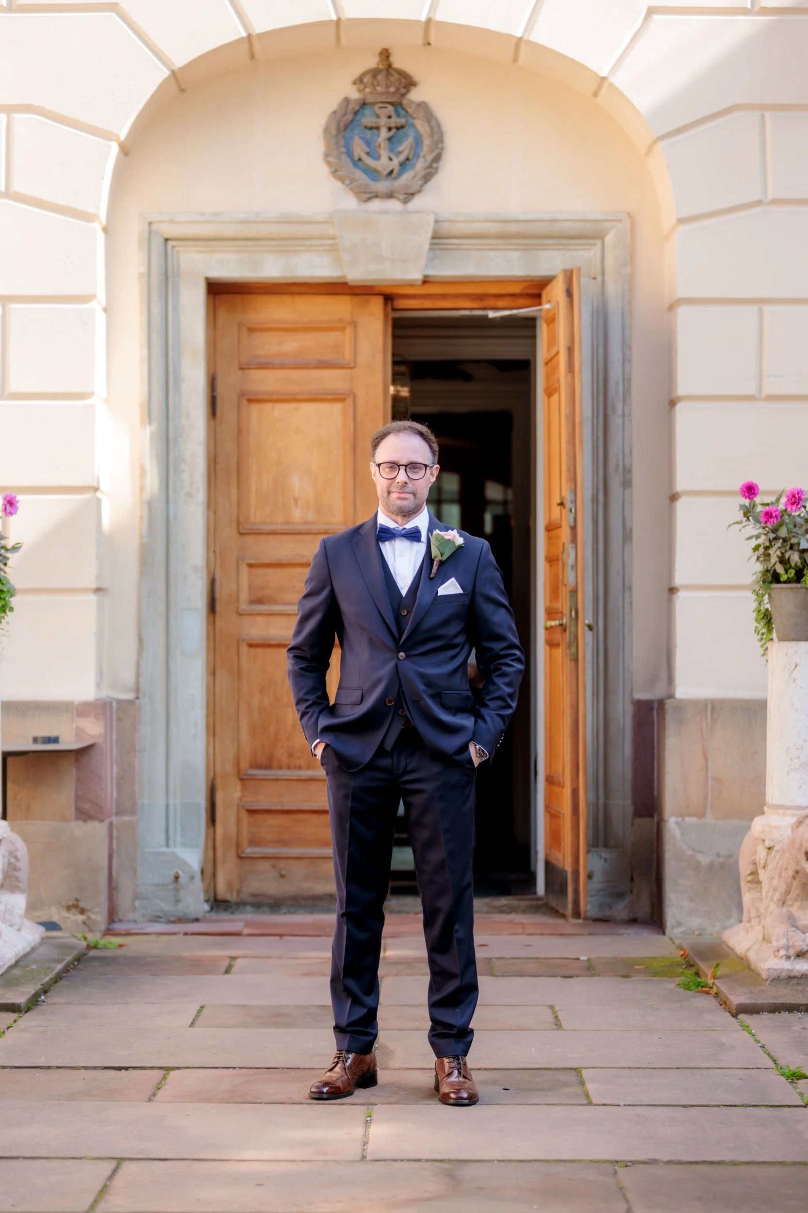 En man i mörk kostym och slips står utanför en byggnad med en öppen dörr. Det finns blommor i krukor på sidorna av dörren. Mannen har glasögon och korsar armarna.