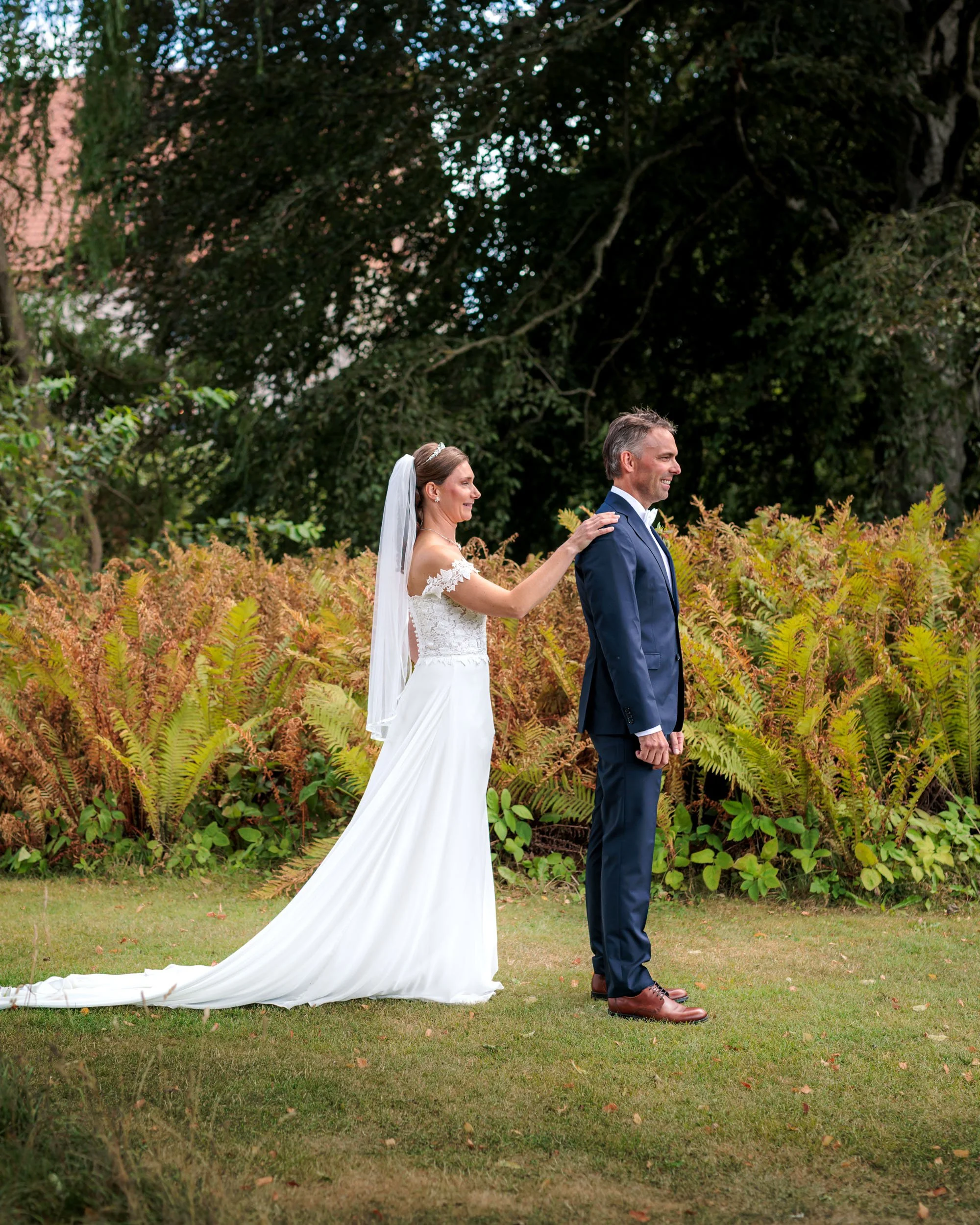 Brudparet står utomhus på en gräsmatta med gröna och bruna växter i bakgrunden, bruden i vit brudklänning och slöja rör vid brudgummens axel, som är klädd i mörkblå kostym.