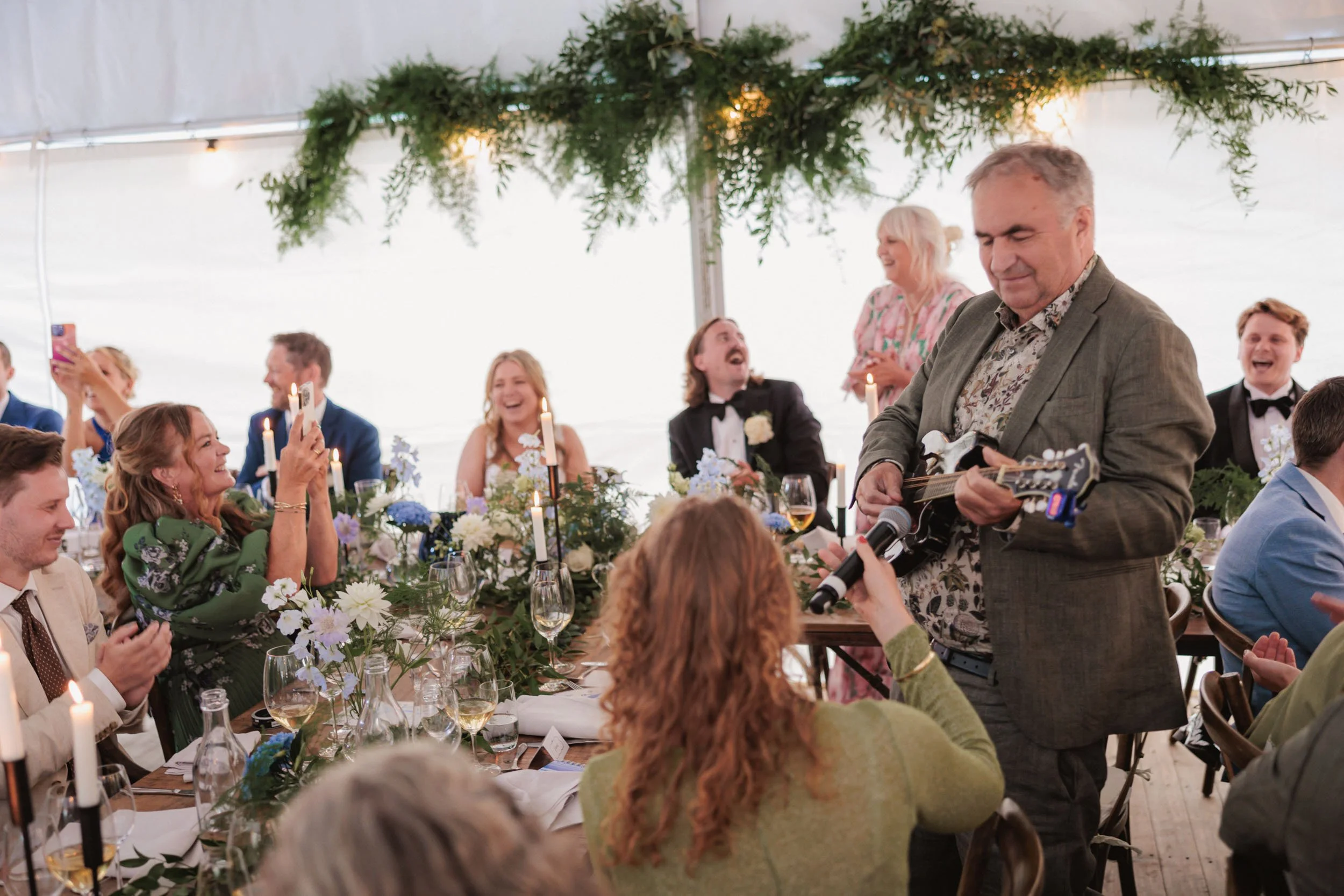 Person spelar gitarr för ett glatt sällskap vid ett bröllop eller fest under en tältat tent med dekoration av gröna växter och ljus.