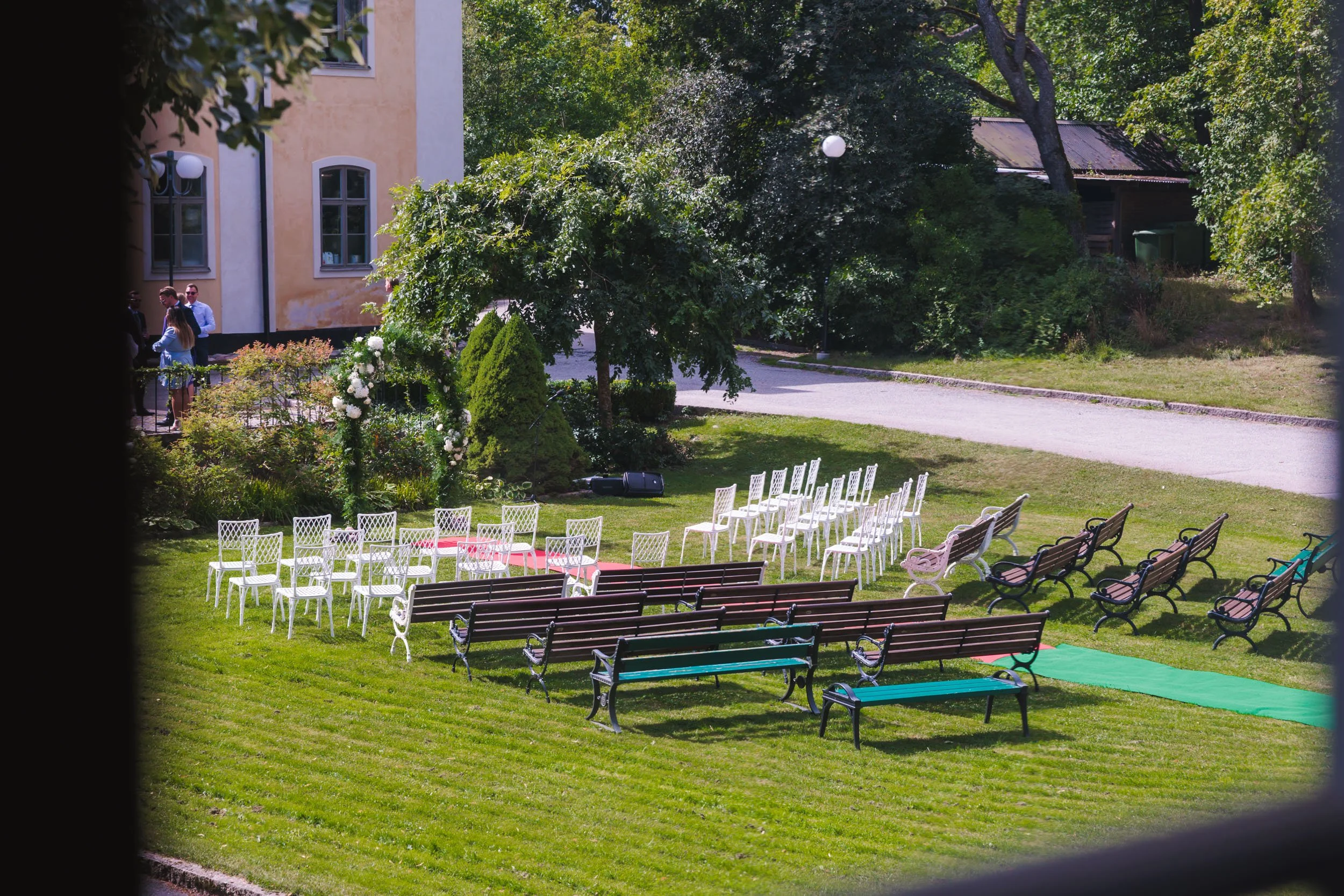 Utsikt över en gräsmatta med rad av vita stolar, andra bänkar och en gångväg, omgiven av träd och växter, förberedd för en utomhusceremoni eller bröllop.