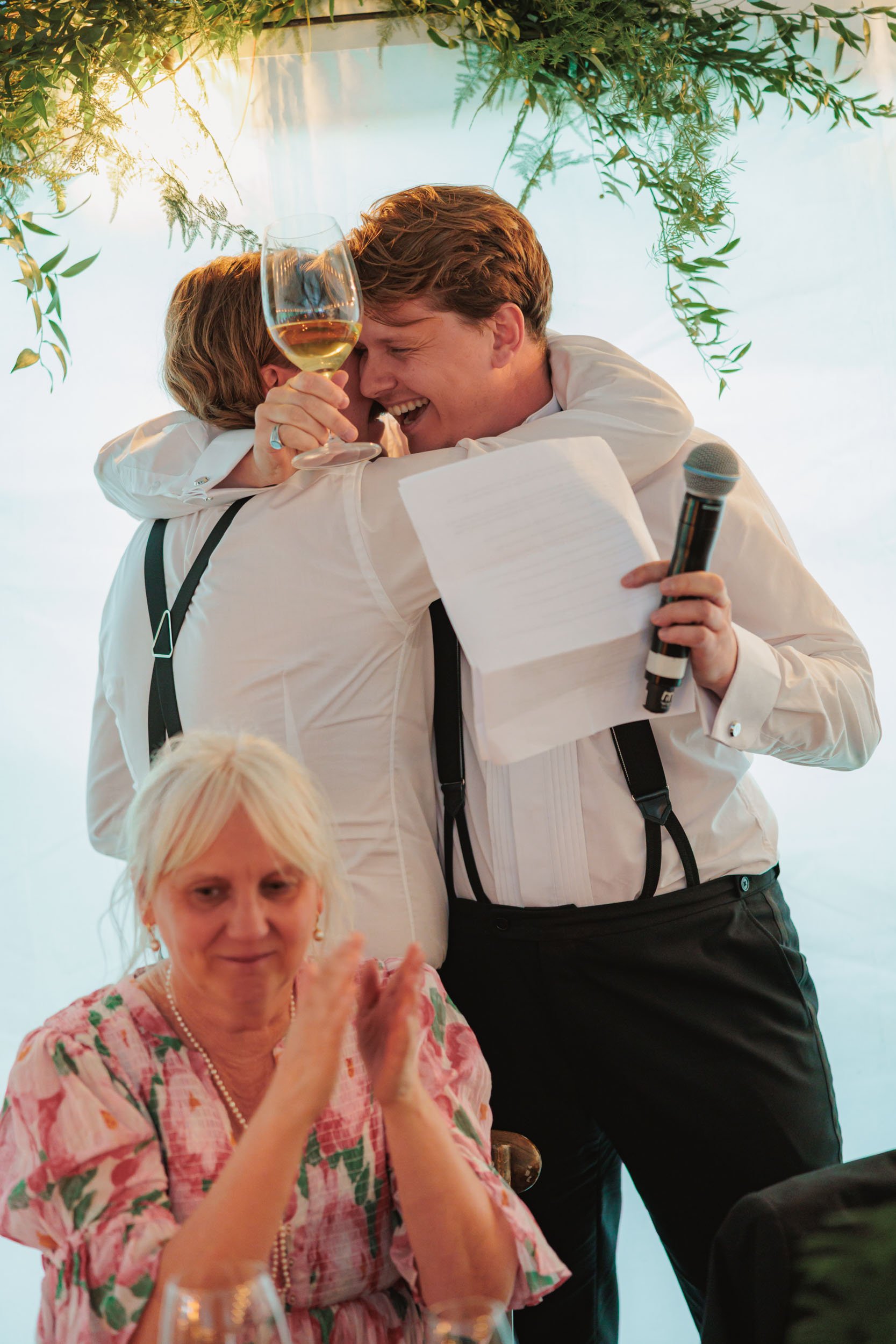 En man och en ung pojke omfamnar varandra i ett firande, pojken håller ett glas vin, den äldre kvinnan sitter framför och applåderar.