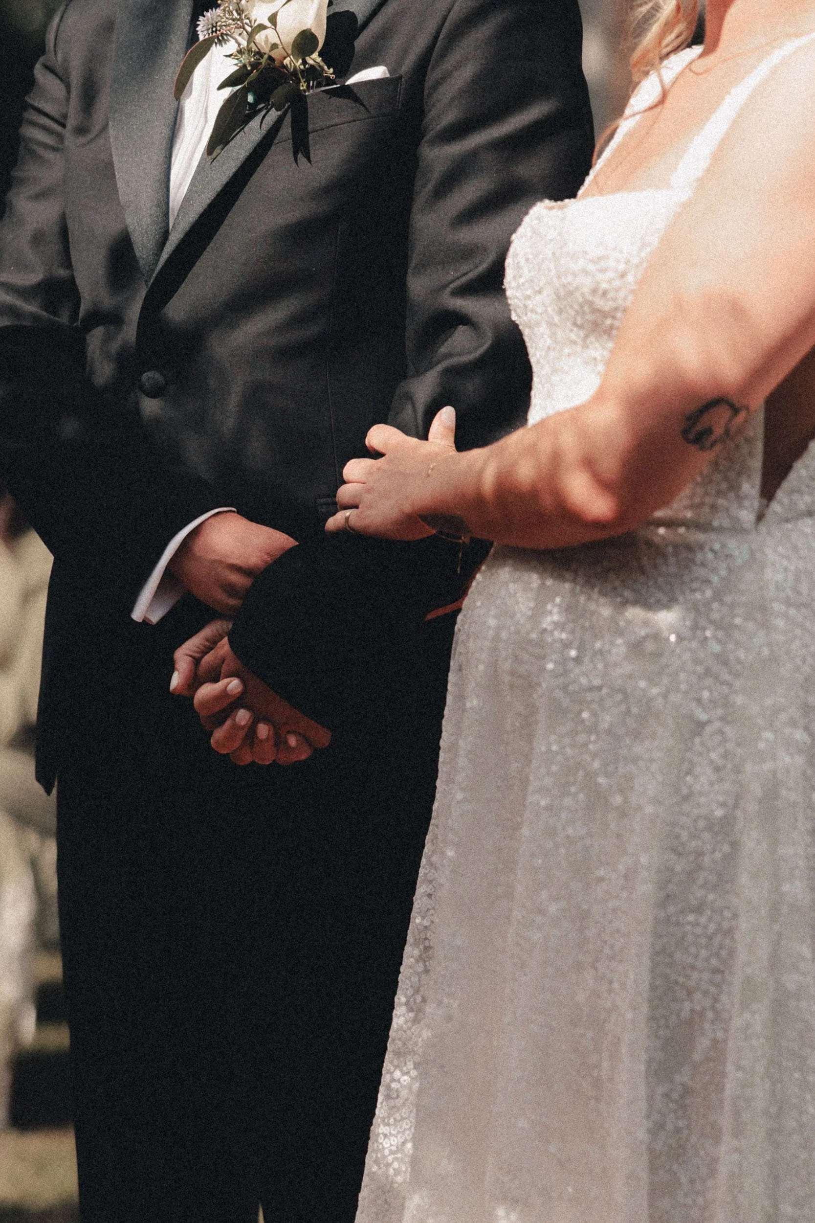 Två personer, en man och en kvinna, håller varandra i handen på ett bröllop. Mannen är klädd i en mörk kostym med vit skjorta, och kvinnan sitter i en vit, glitterklädd brudklänning.