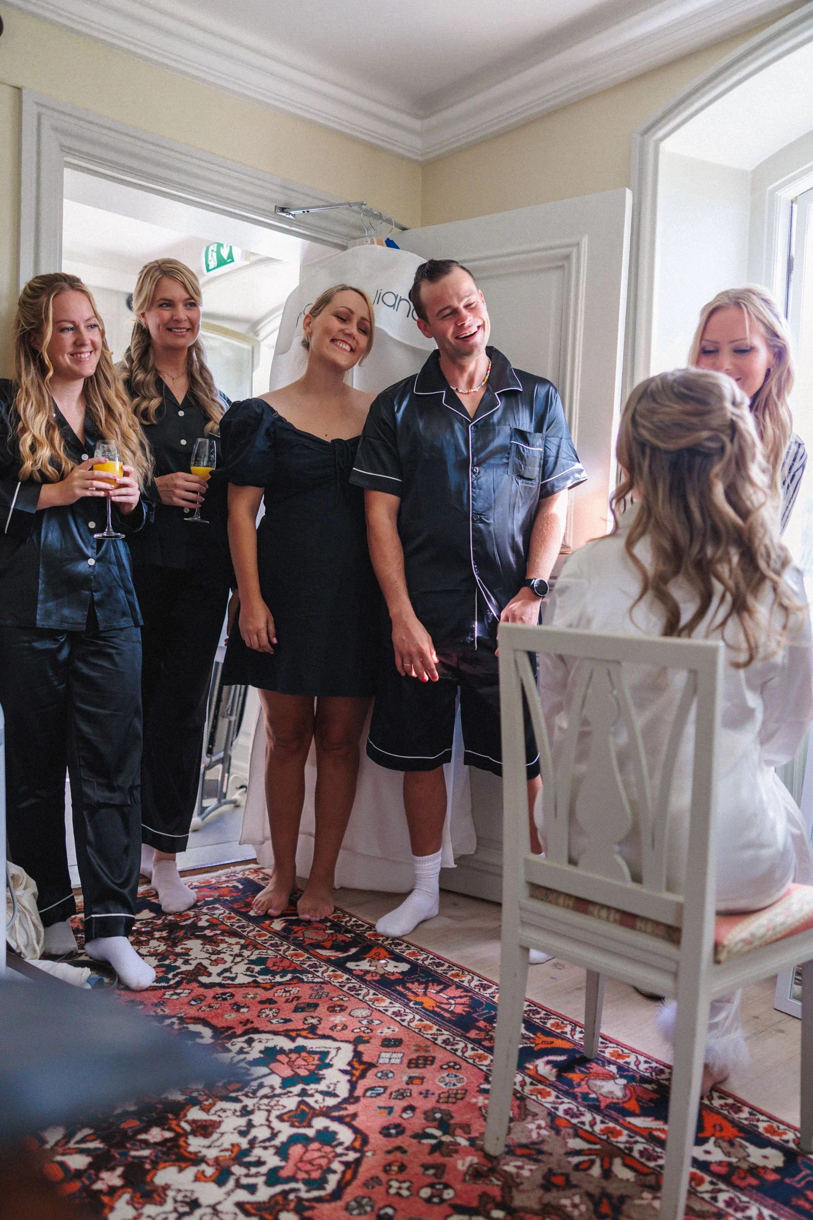 En grupp människor i pyjamas som ler och skrattar, troligen under ett bröllopsförberedelse eller morgonsamling, i ett ljust rum med en matta.