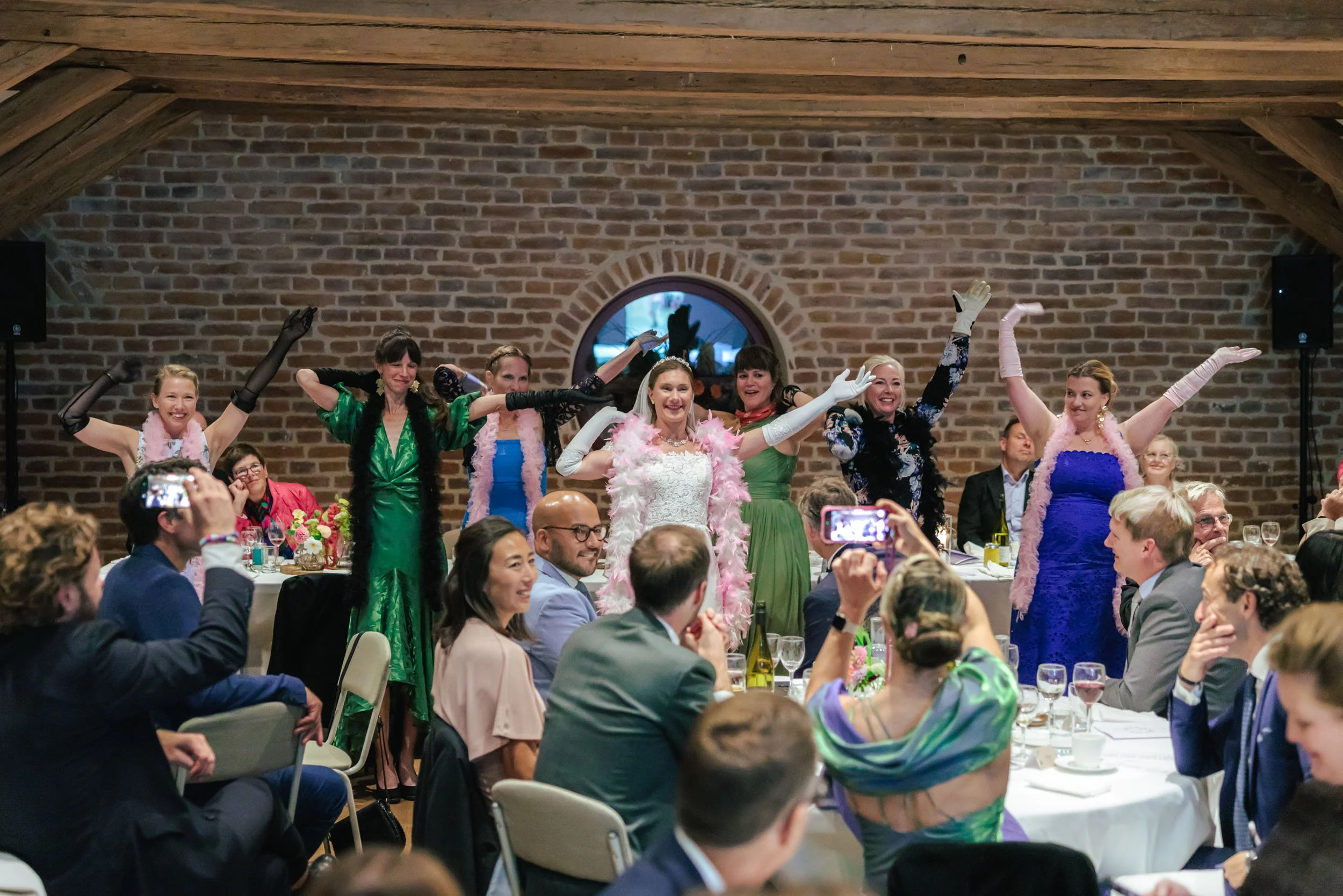Grupp av kvinnor och män som dansar på ett bröllop, med ett brudpar i mitten, i ett rustikt rum med tegelväggar.
