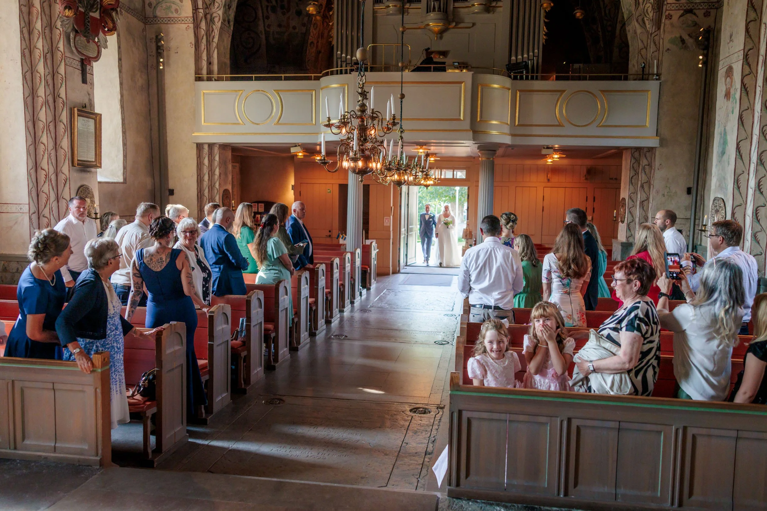 Människor samlade i en kyrka för ett bröllop, kvinnor och män står i rader, några fotograferar och ser glada ut, ett par promenerar in genom dörren i bakgrunden.