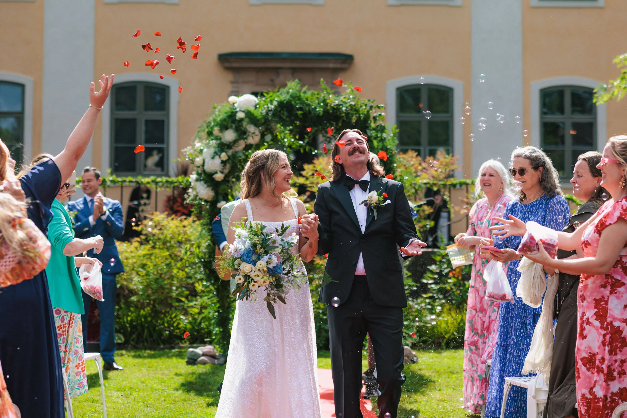 Bröllopspar går ut efter vigsel omgivna av gäster som kastar blomblad – utomhusbröllop i Sverige