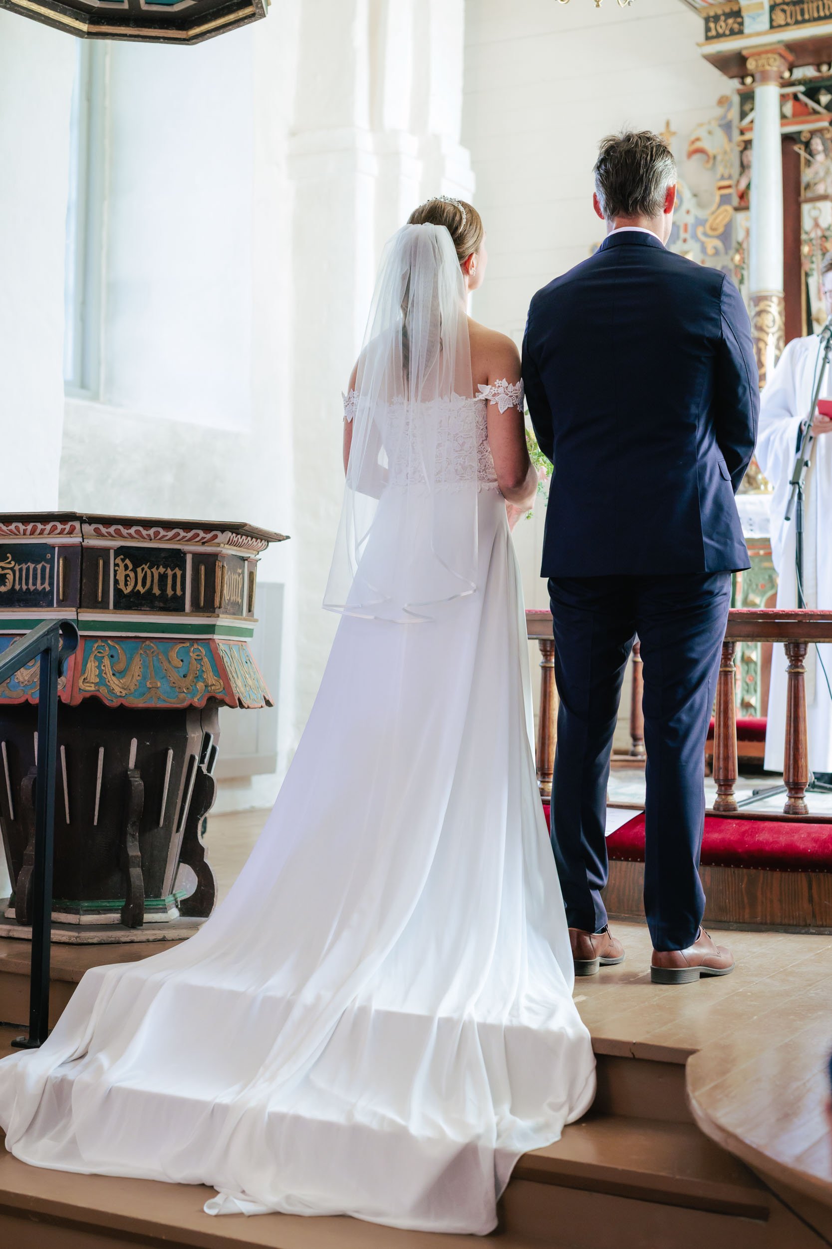 Brud och brudgum står vid altaret under ett bröllop i en kyrka. Bruden bär en vit vigselklänning med dragkedja och ett tiarliknande hårband, samt en slöja. Brudgummen är klädd i en mörkblå kostym och ljusbruna skor.