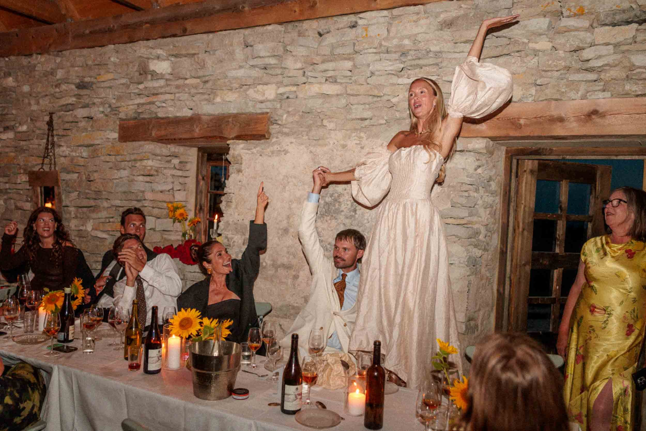 En grupp människor firar ett bröllop, en kvinna i bröllopsklänning står på en stol och verkar vara drottningen av festen medan andra skrattar och pekar mot henne. Det är mysigt belyst med ljus, i ett rum med stenväggar.
