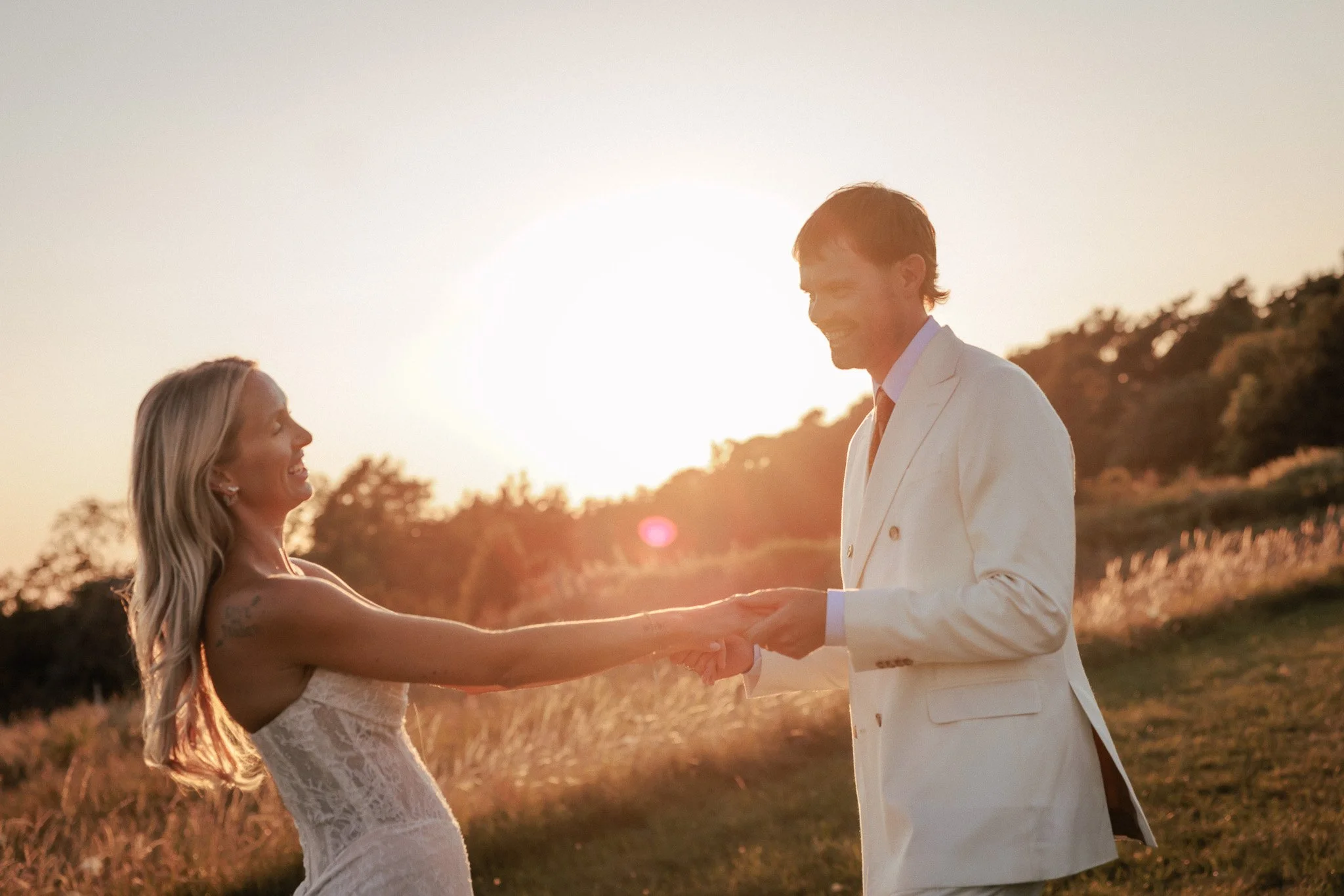 Brudpar håller händer och snurrar i öppet fält med stark solnedgång och linsflare i bakgrunden