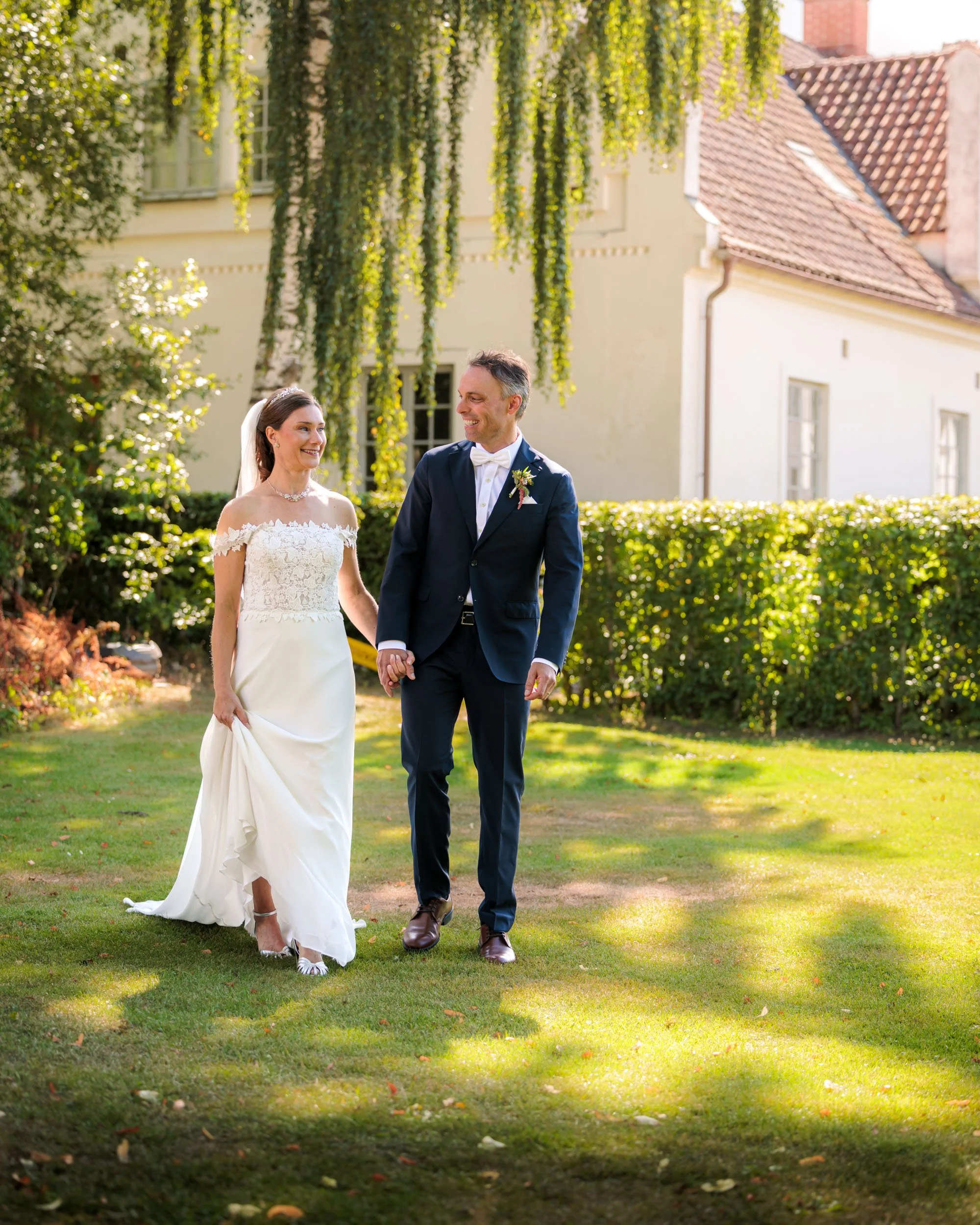 En nygift brud och brudgummi går hand i hand i en trädgård på en solig dag.