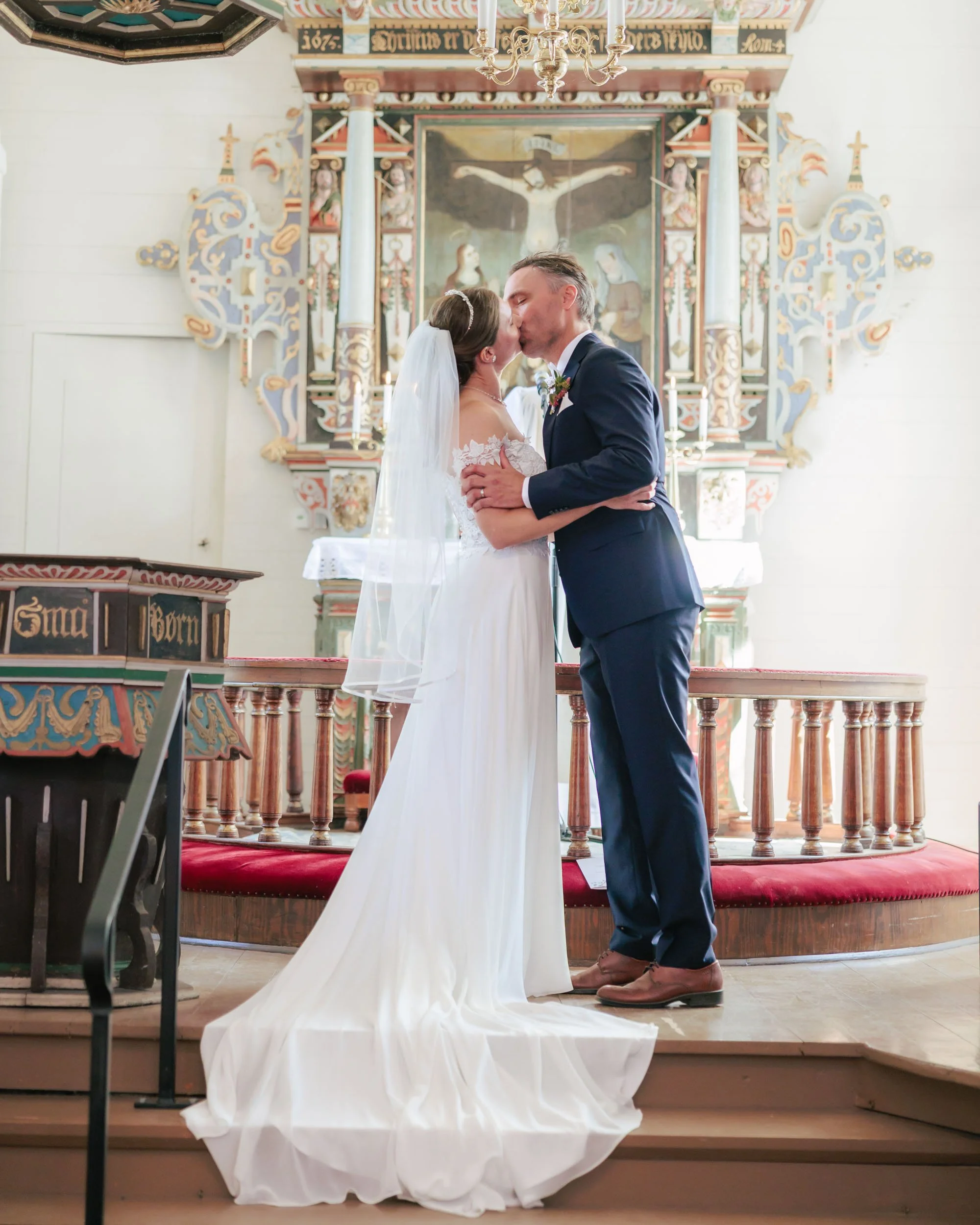 Nygifta par kysser i en kyrka, med en altartavla och snirkliga dekorationer i bakgrunden.