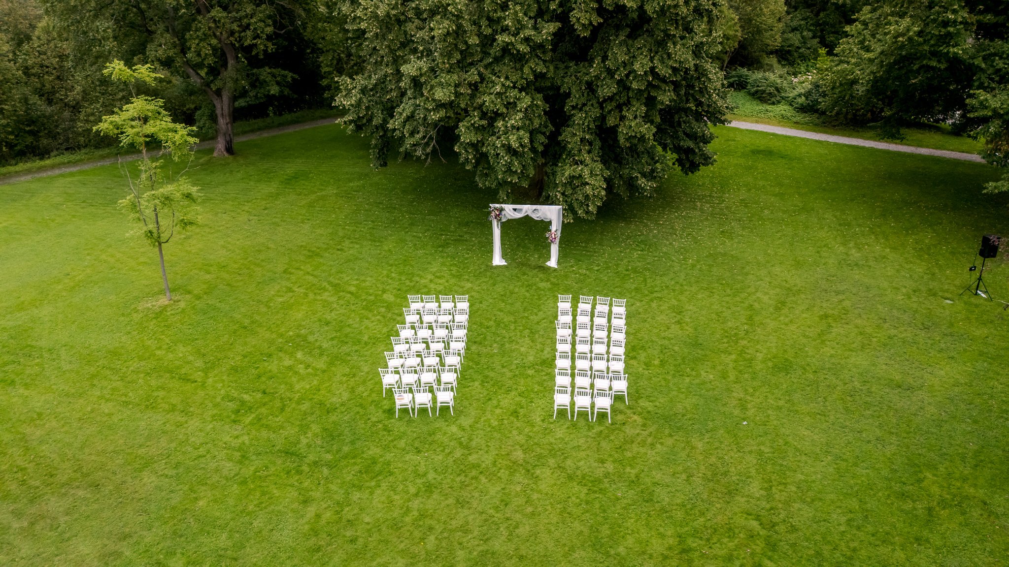 Drönarbild över tom ceremoniplats med vita chiavari-stolar i rader och vit draperibåge på grön gräsmatta
