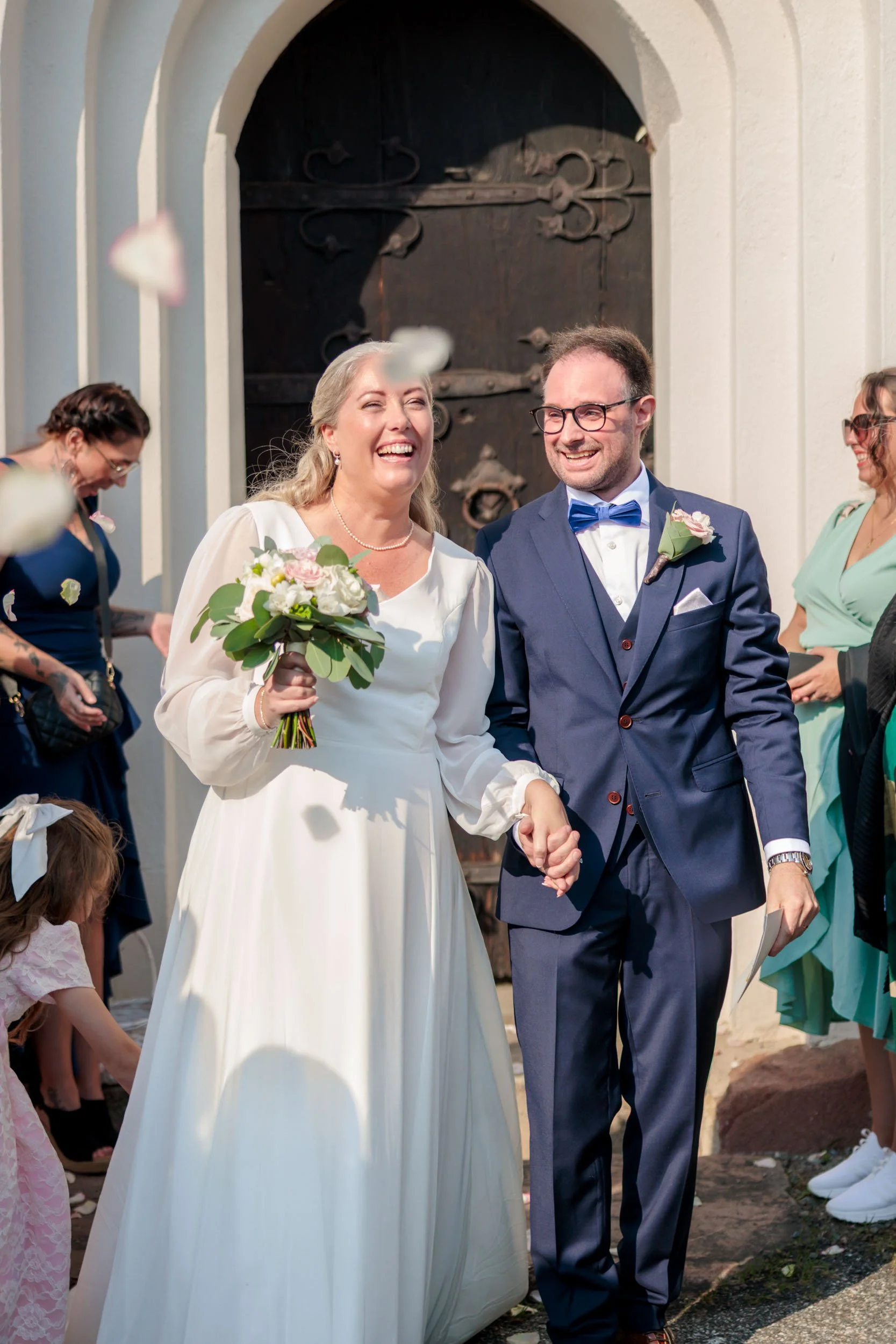 Ny gift kvinna och man håller handen och ler utomhus framför en stor mörk träport, omgiven av vänner och familj.