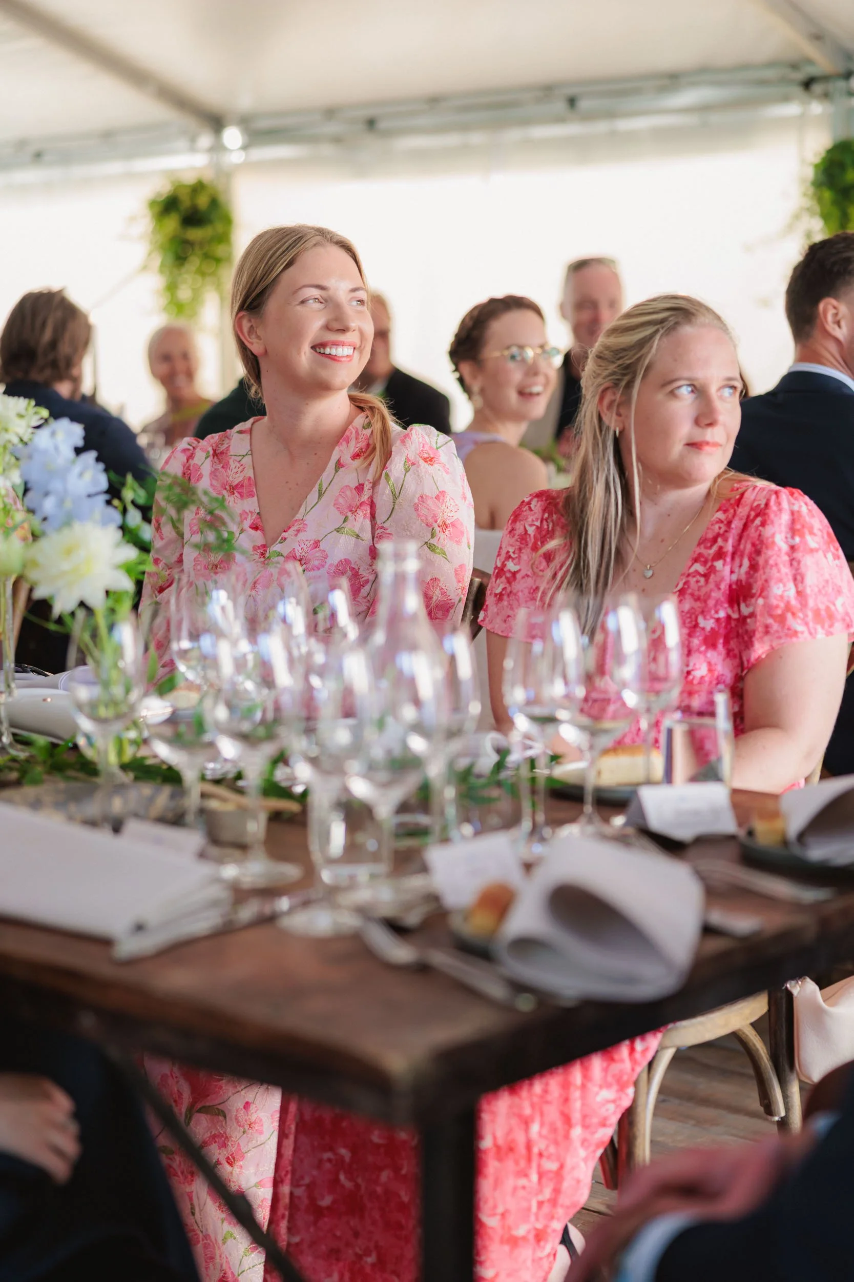 Flera personer sitter vid ett bord under en festlig tillställning, där två kvinnor i fyrkantiga mönstrade klänningar är i fokus. Den ena kvinnan ler brett, medan den andra tittar bort. På bordet finns tomma glas, servetter och blomsterarrangemang.