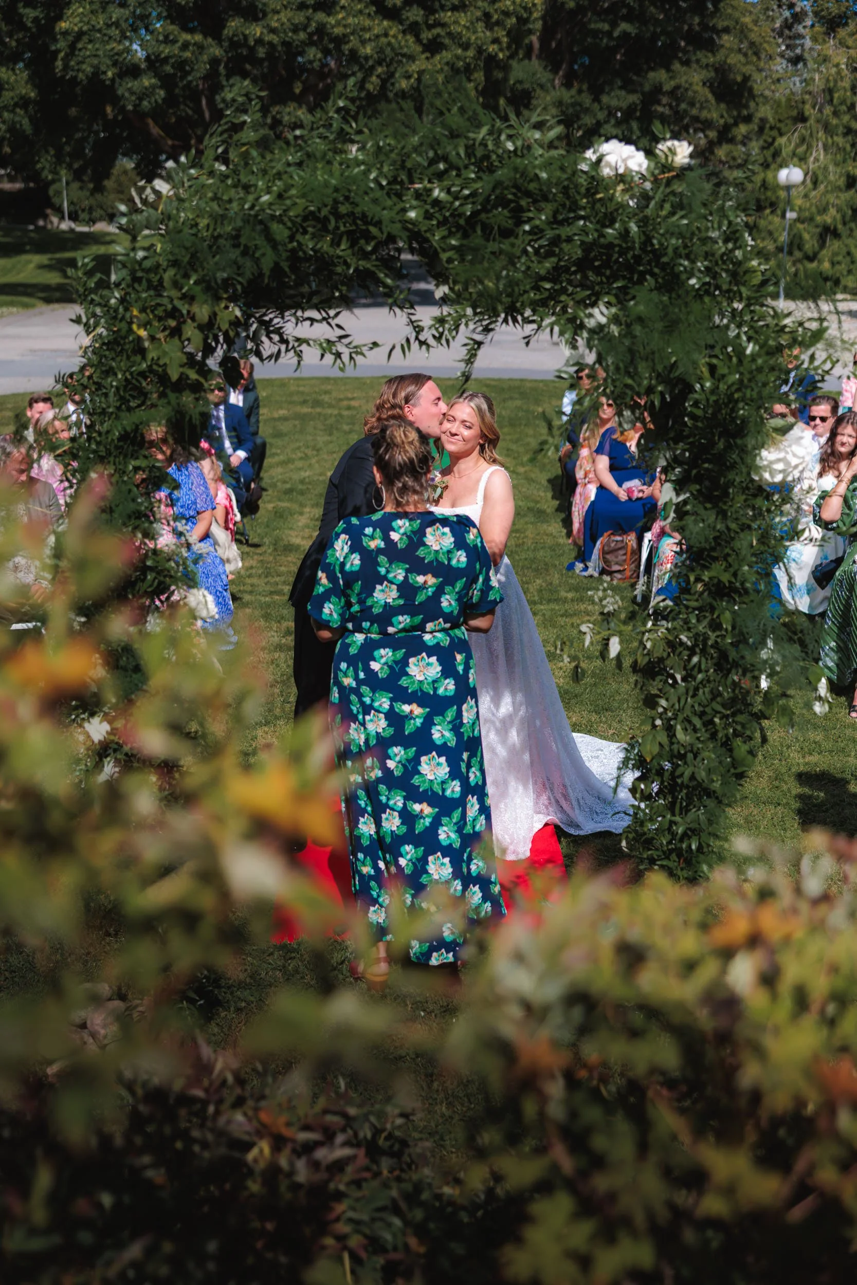 Brudpar står under ett blomsterarrangemang, omgiven av gäster, på en gräsmatta under en utomhusceremoni.