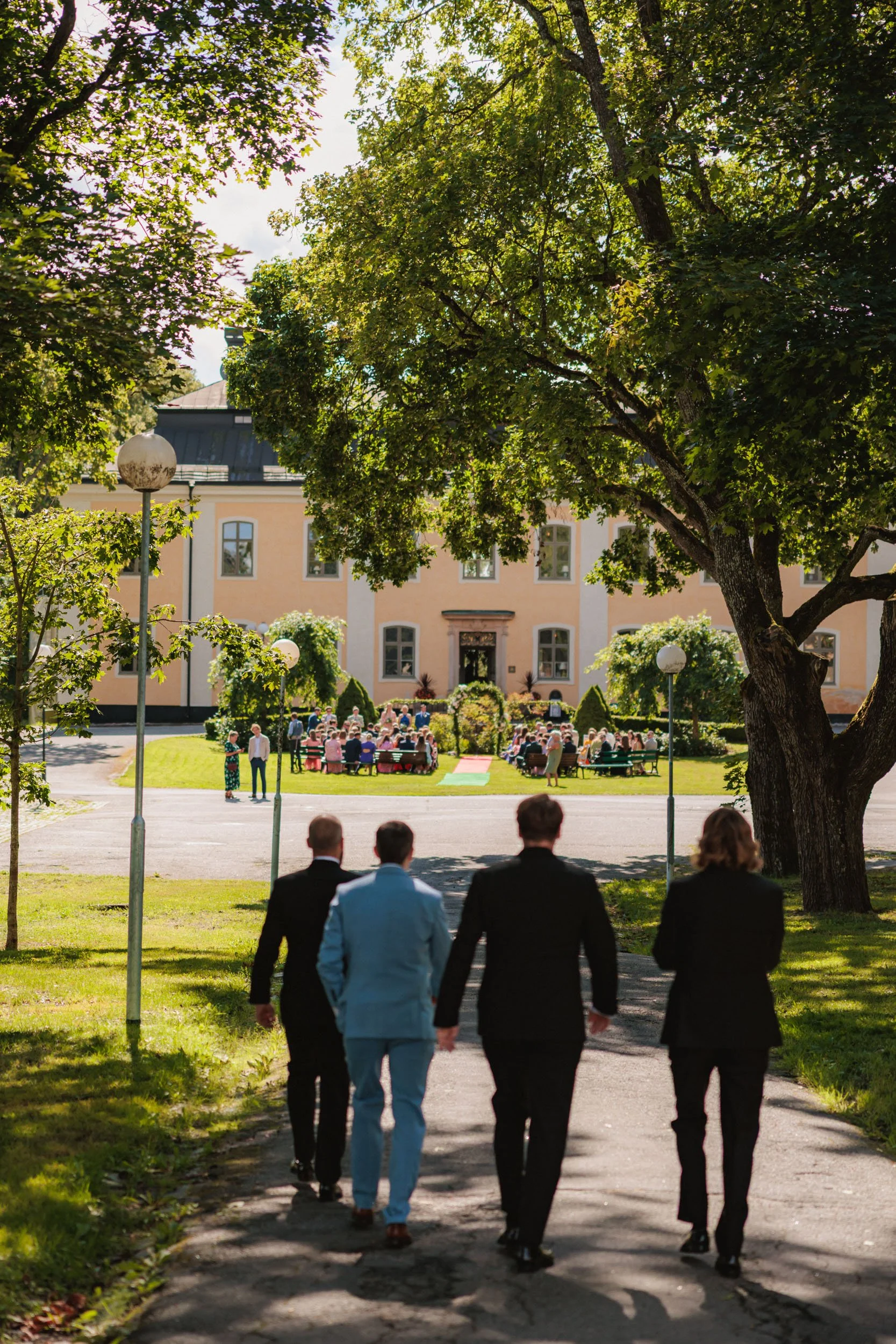 Människor i formella kläder går mot en utomhusfest framför en stor byggnad, som är omgiven av gröna träd och en gräsmatta.