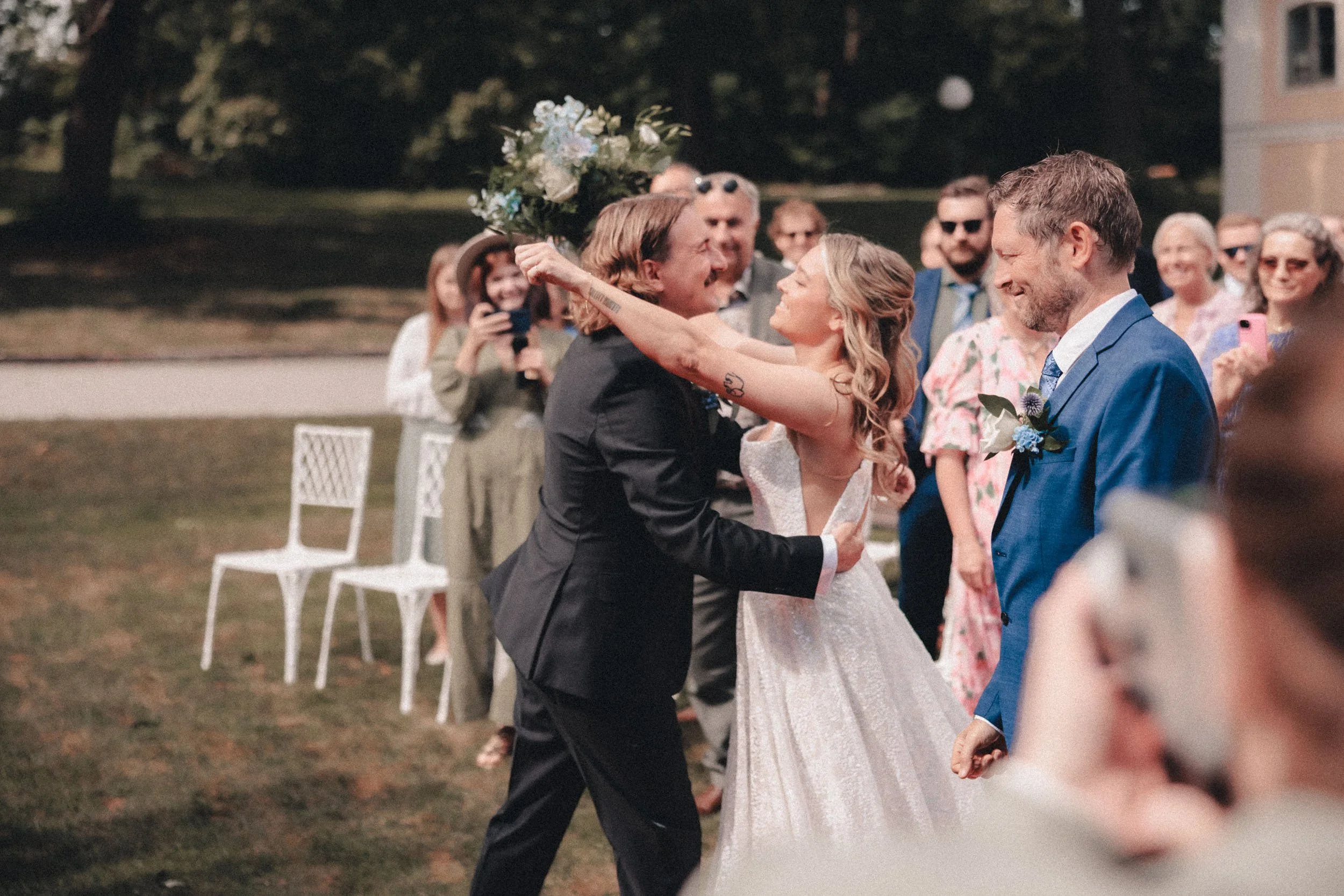 En bröllopsdans på ett utomhusbröllop, med en man och en kvinna i centrum som ler och håller varandra, omgivna av gäster som fotograferar och iakttar dem.