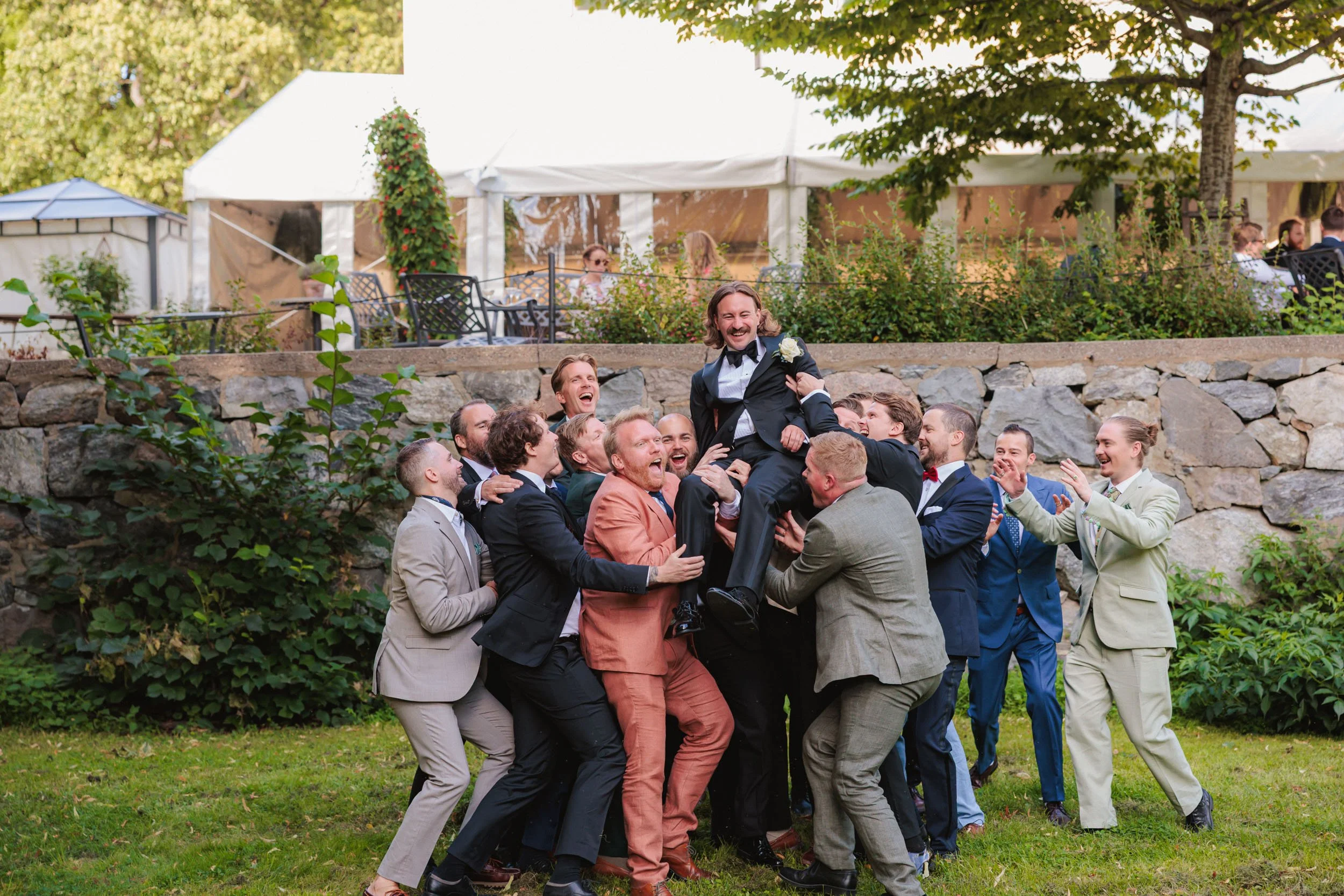 Grupp bröllopsgäster som lyfter upp en man i kostym, skrattande utomhus.