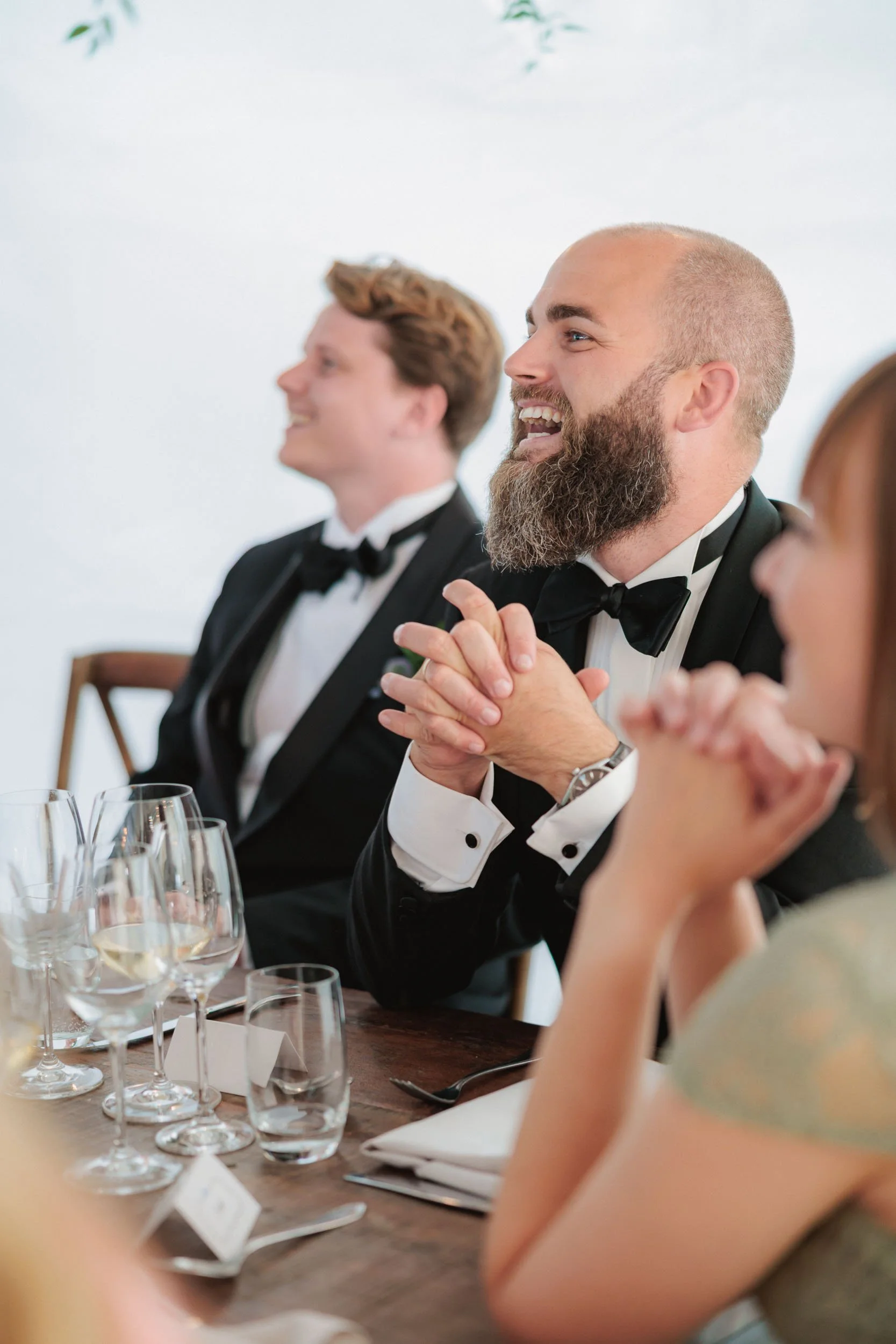 Män i formella kläder som skrattar och ler vid bord med vinglas under en fest eller bröllop.