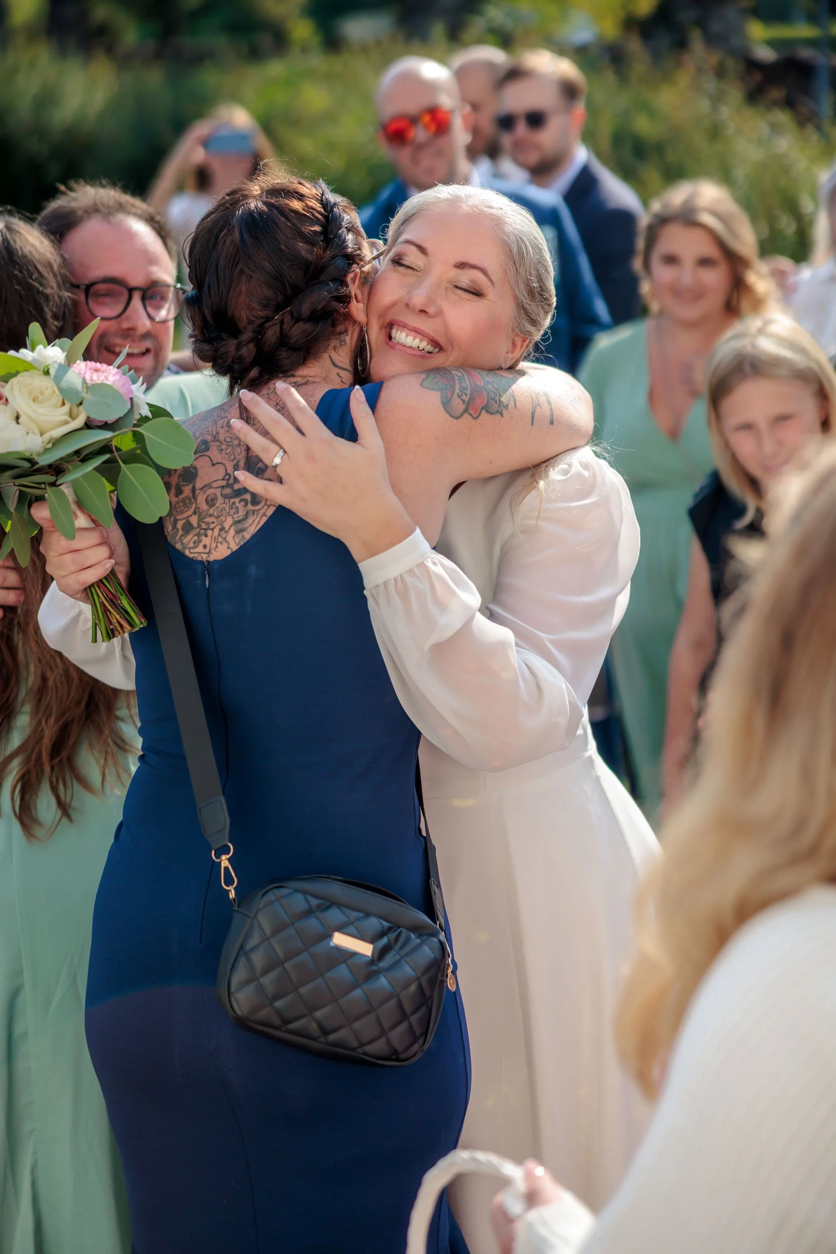 Två kvinnor kramas glatt på ett utomhusbröllop, omgivna av glada gäster.