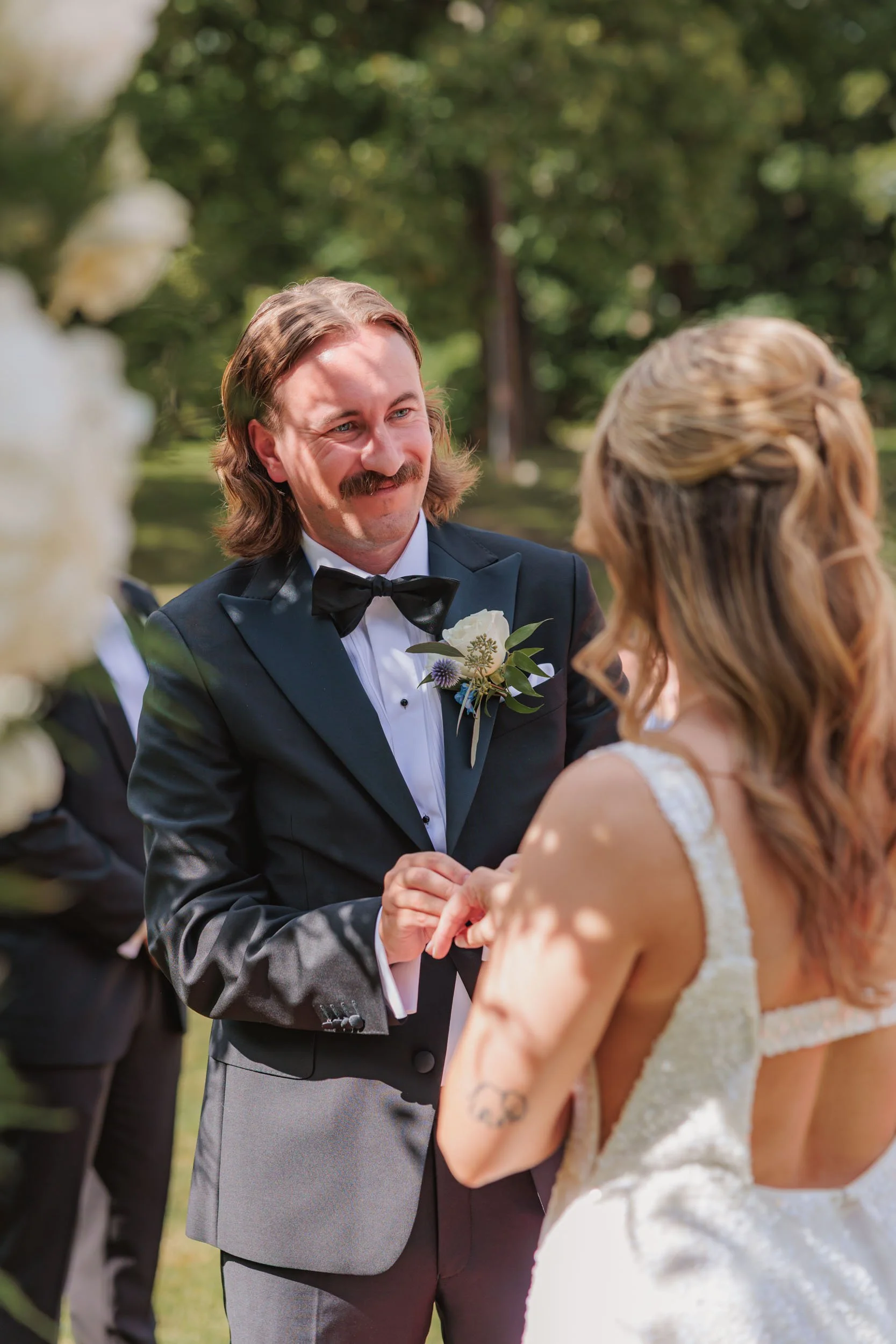 En man i svart frack med fluga håller handen på en kvinna i vit brudklänning under ett utomhusbröllop, lummig grön bakgrund.