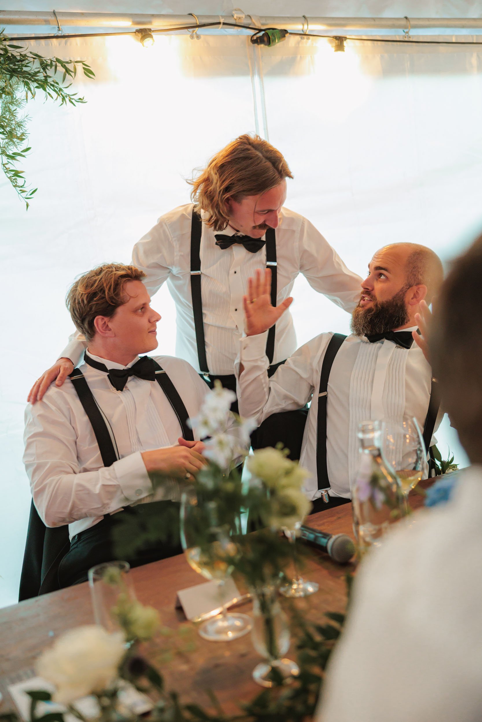 Tre män i vita skjortor, svarta fluga och svarta hängslen, samt en man i samma klädsel, interagerar under ett festligt tillfälle. De sitter vid ett bord med blomsterarrangemang, vattenglas och mikrofon. En man reser sig och ger handtecken till en ann