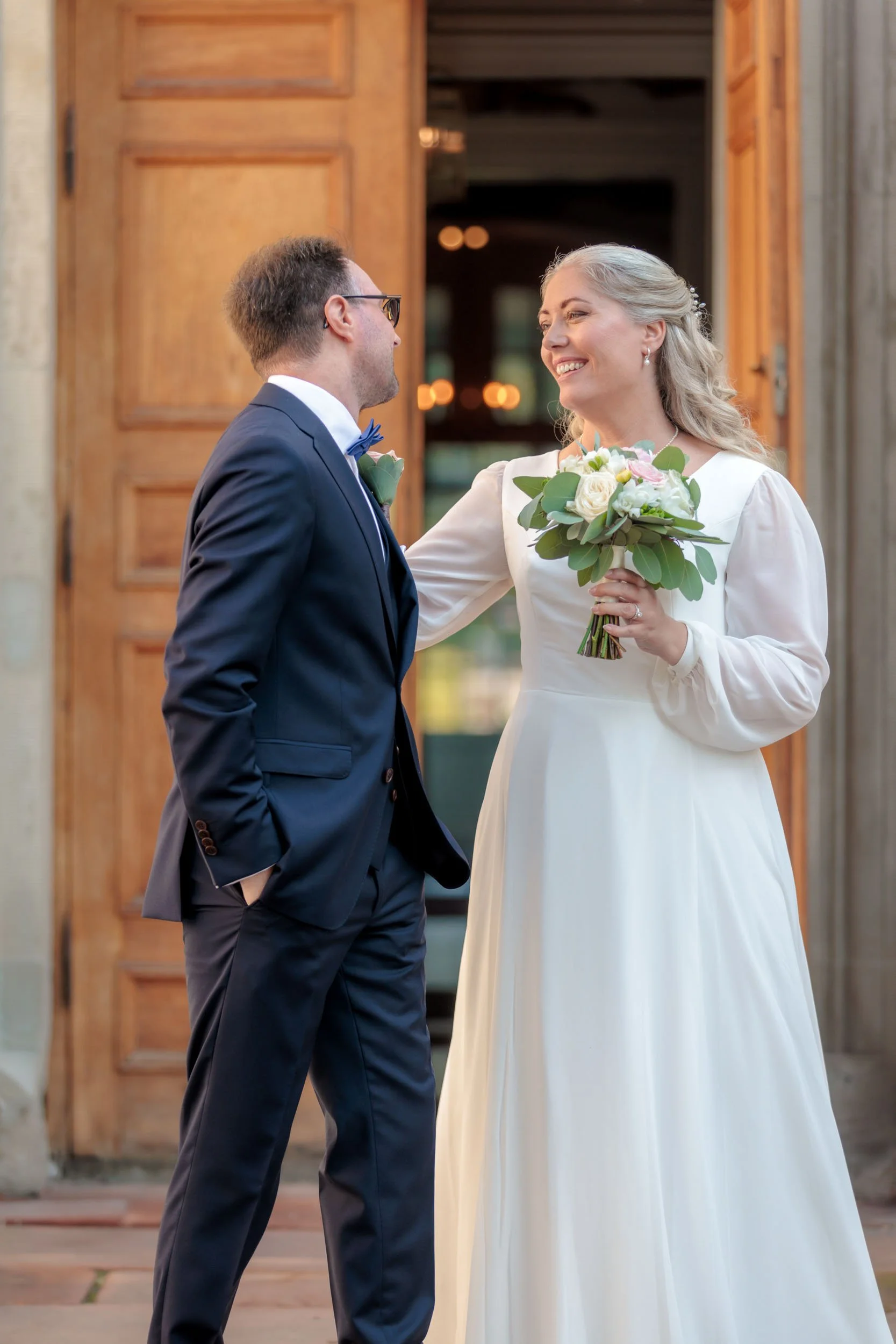 En brud och en brudgummar som står framför en trähusdörr, ler mot varandra under deras bröllop.