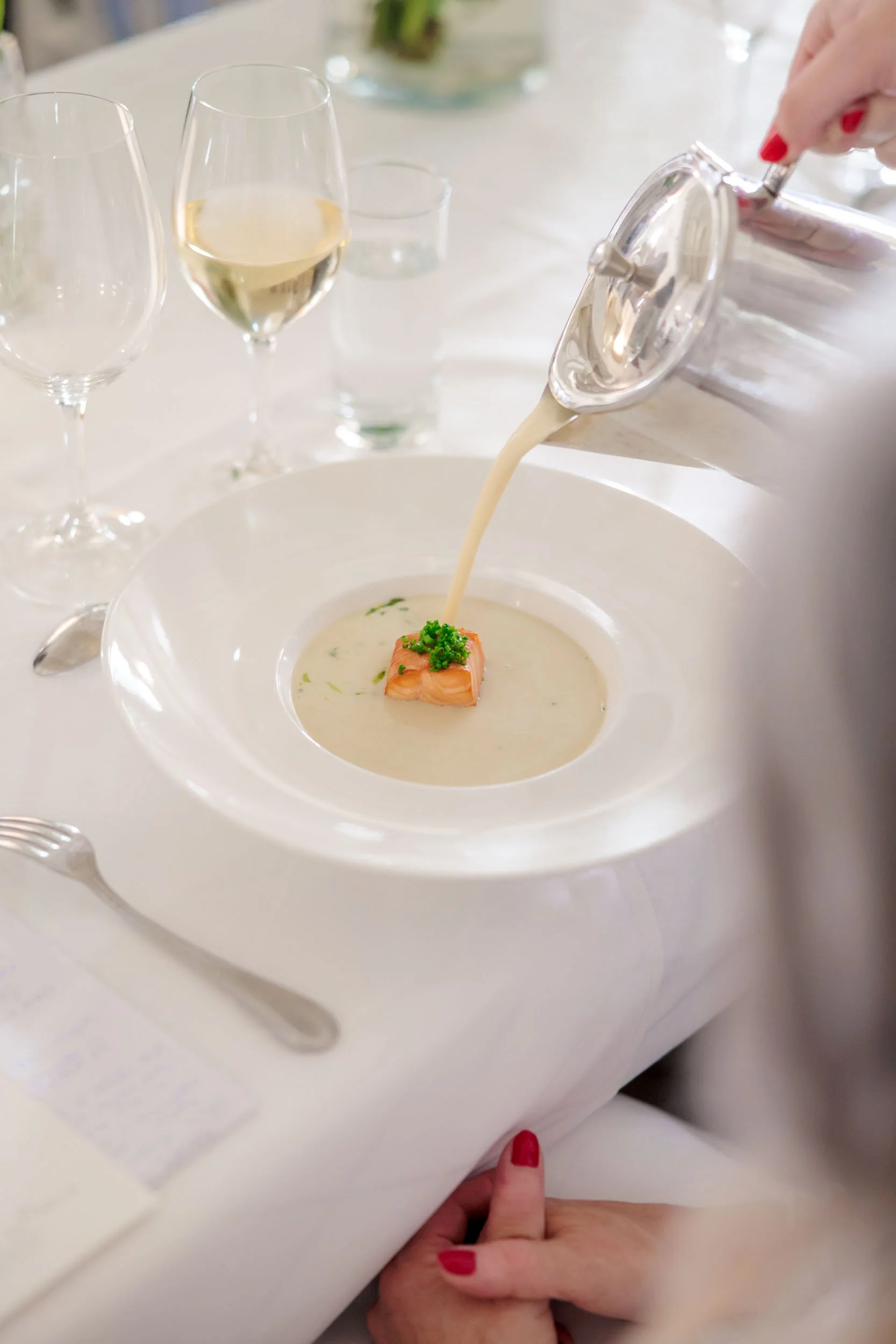 En hand häller en ljus soppa med lax och grönt i en stor vit skål, med snabba glas vin och vatten på ett vitt bord.