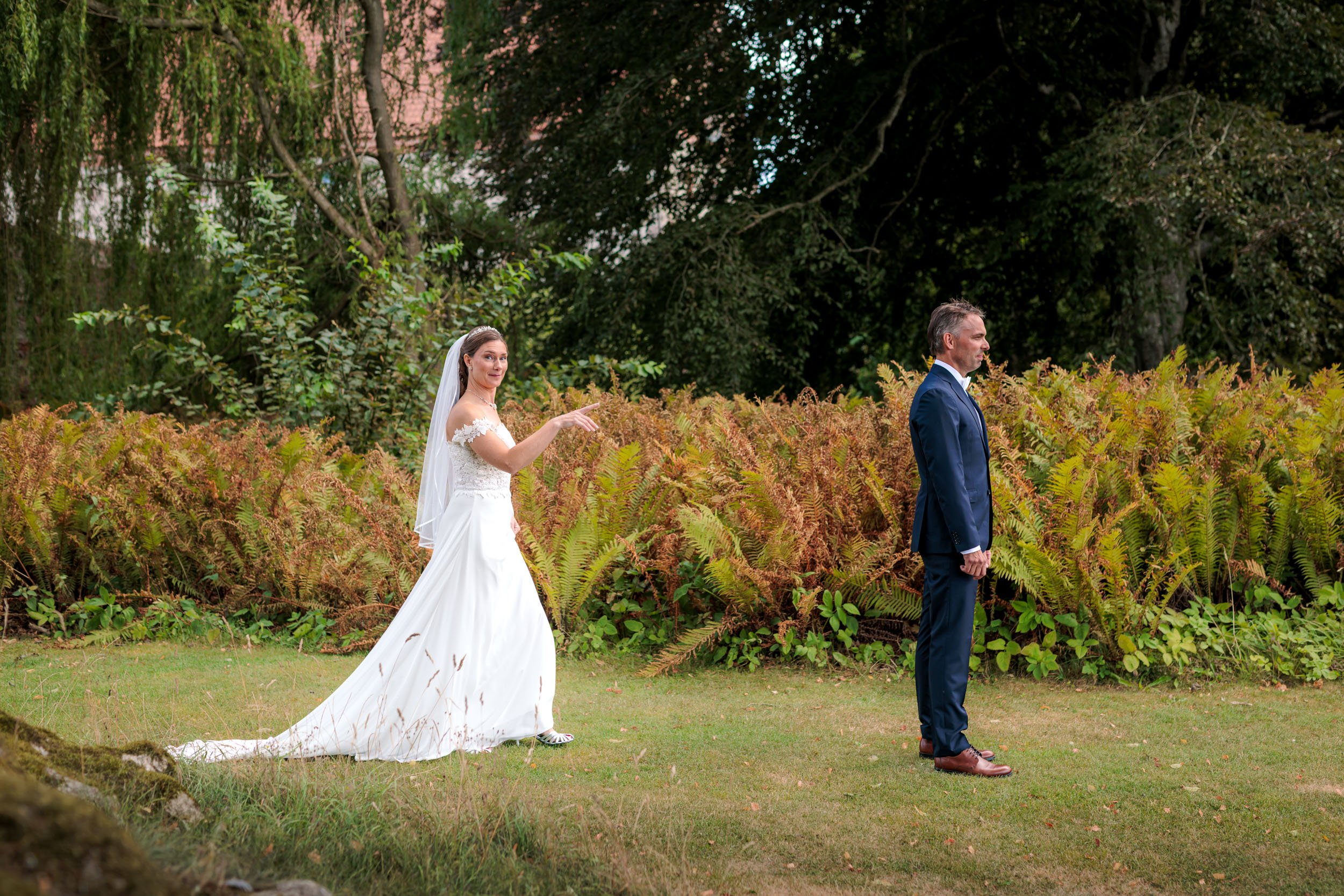Brudgum och brud står på en gräsyta i en trädgård, med bruden som vinkar eller pekar mot gommen.