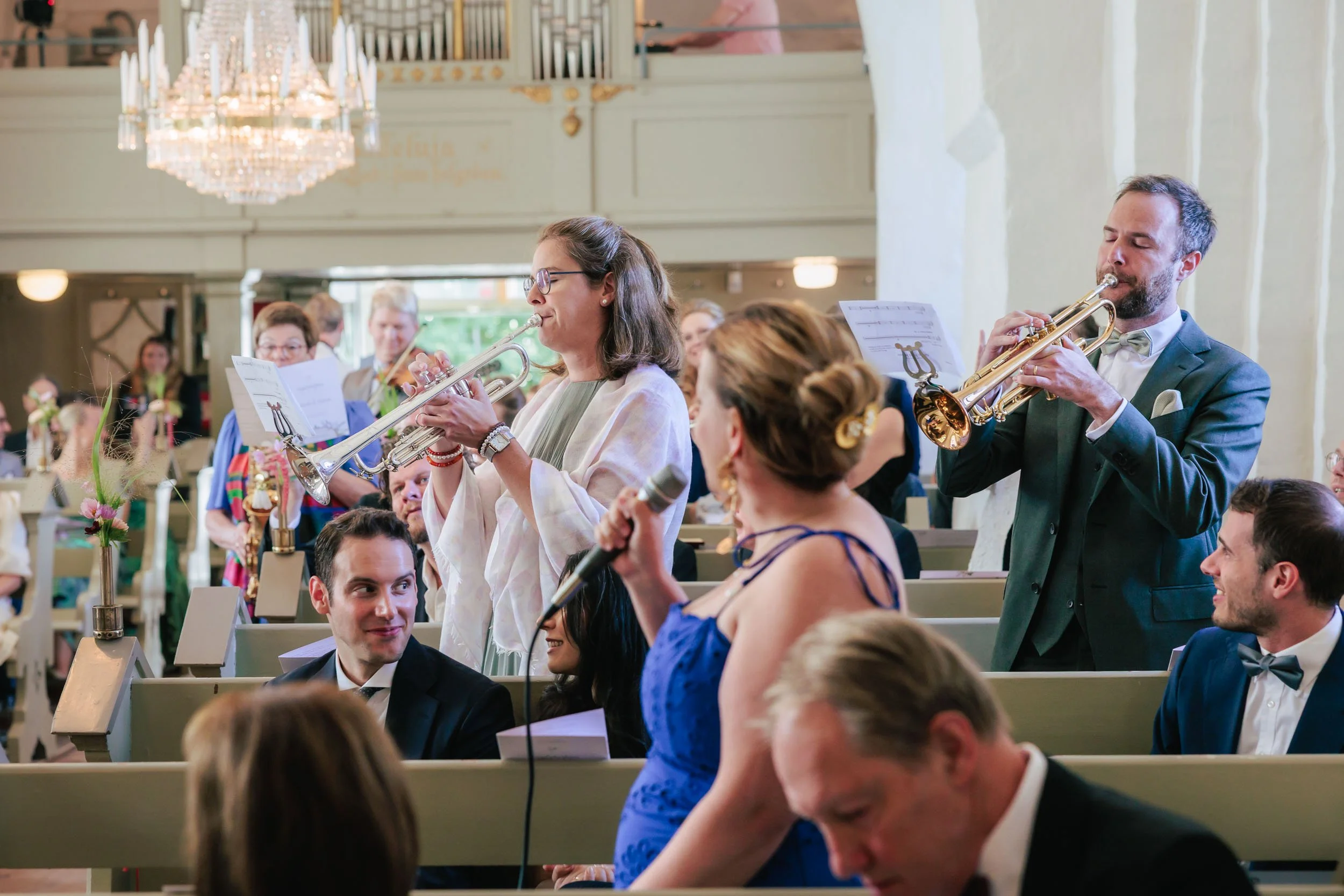 Människor i formella kläder inomhus, som sjunger och spelar instrument, i ett festligt sammanhang, möjligen ett bröllop eller en musikalisk presentation.