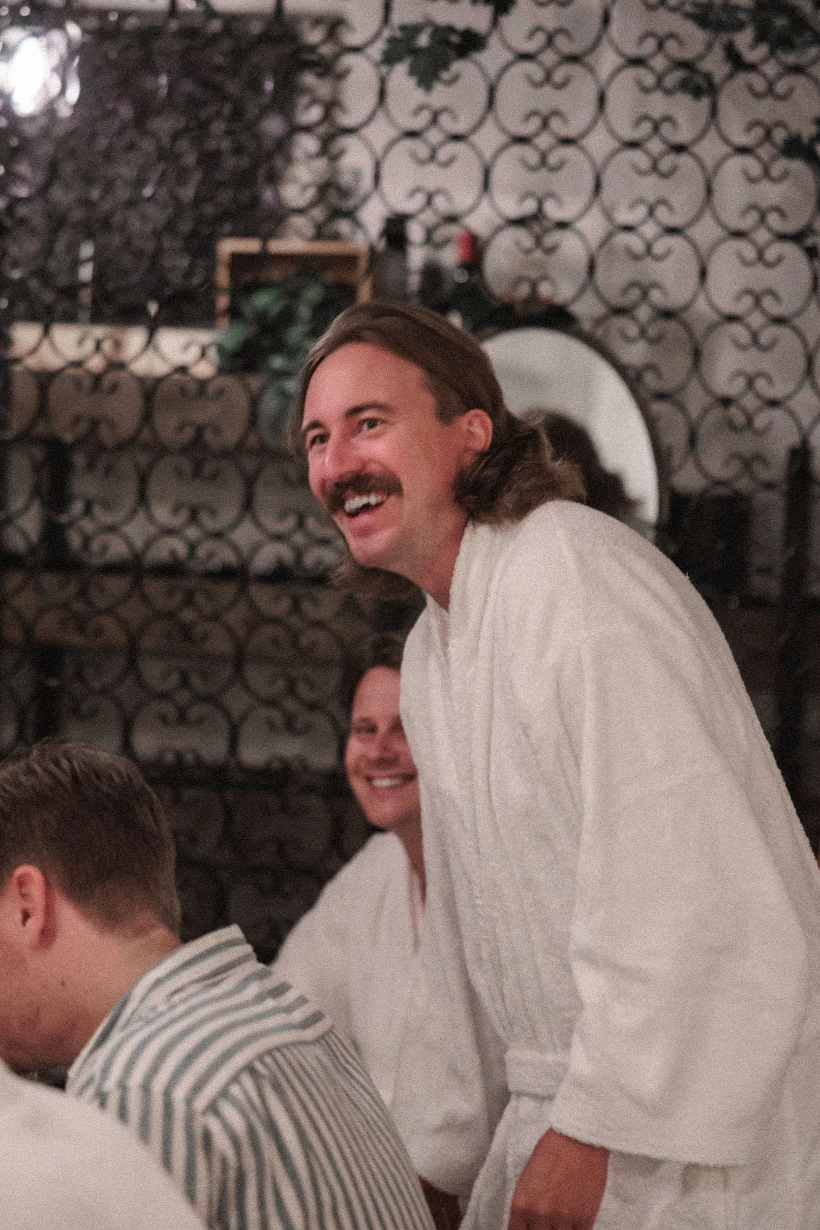 Vänjer ett leende på en man med långt hår och mustasch, alla iklädda vit morgonrock, i ett inomhusmiljö med svart och vitt mönstrad vägg.